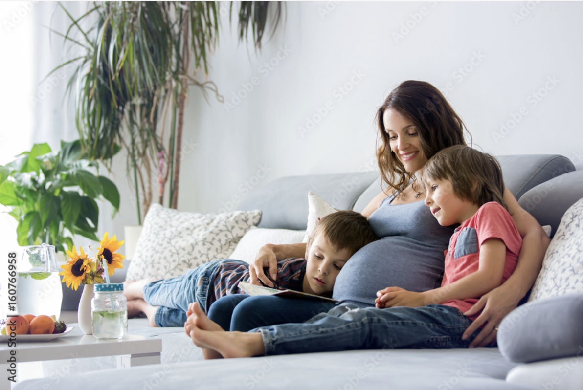 A pregnant woman sitting on a sofa with two young boys, one on each side, in a living room, smiling and looking at a book.