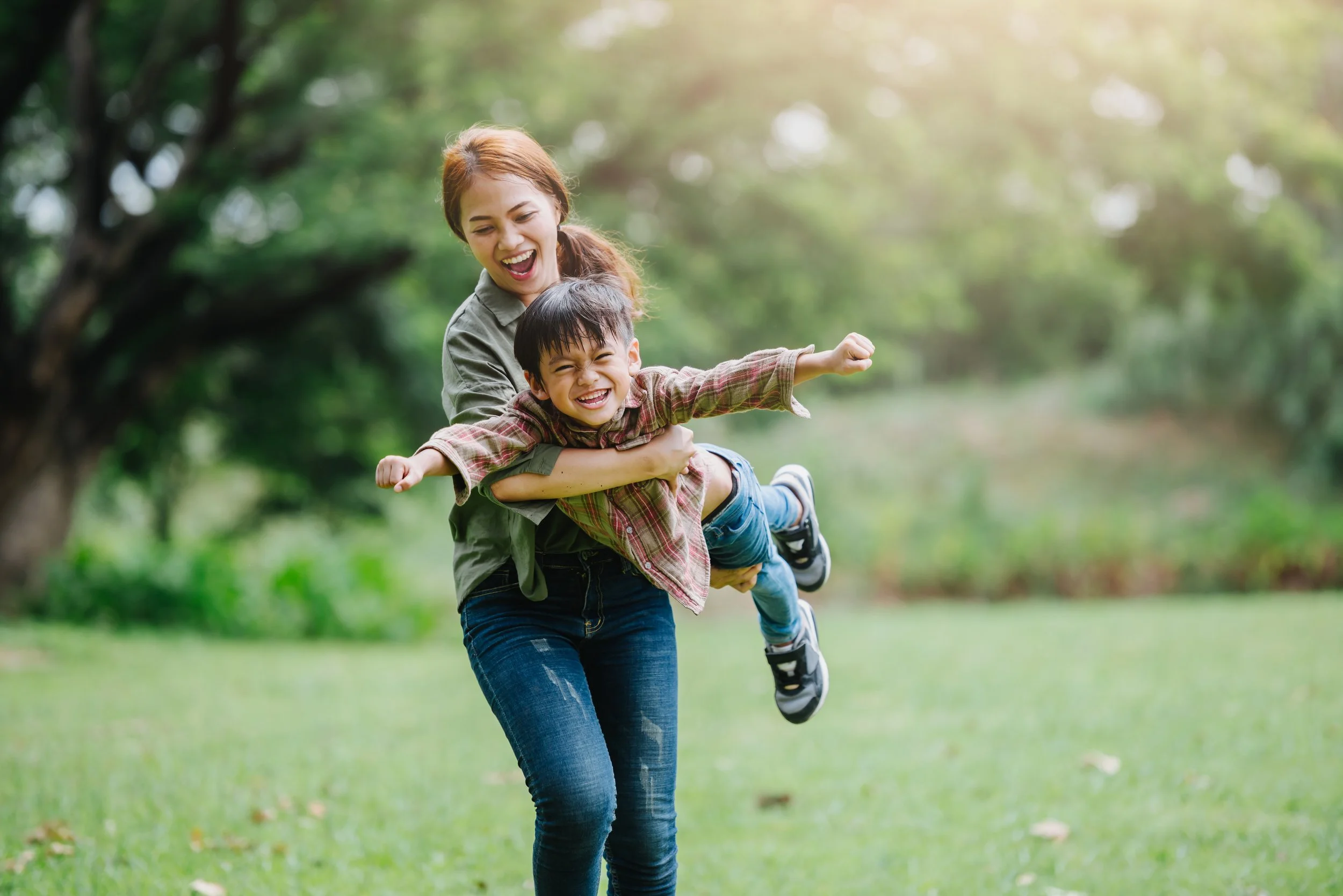 A woman and young boy playing outdoors in a grassy park, with the woman lifting the boy in her arms, both smiling joyfully.