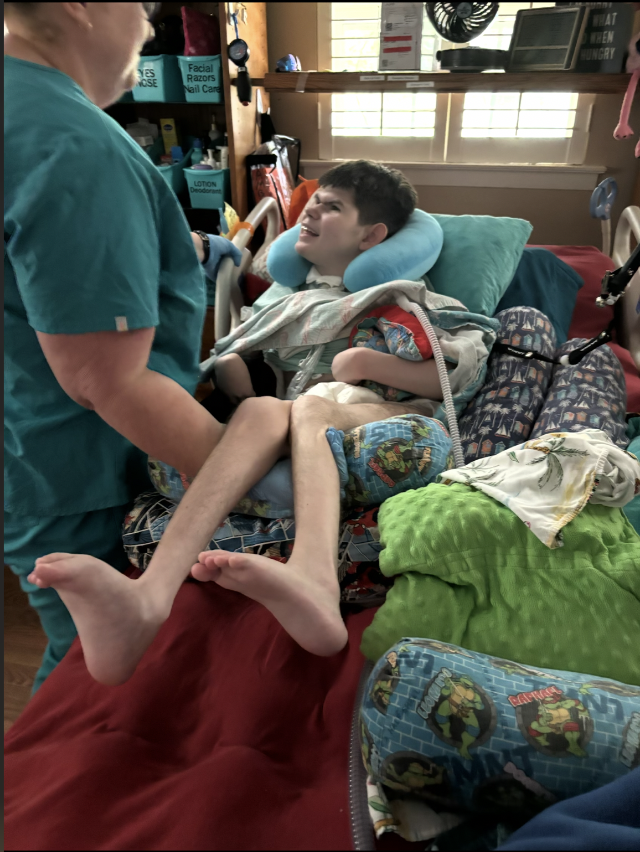 A young boy in a hospital bed with a neck brace, smiling and holding hands with a visitor, in a hospital room with medical supplies and windows in the background.