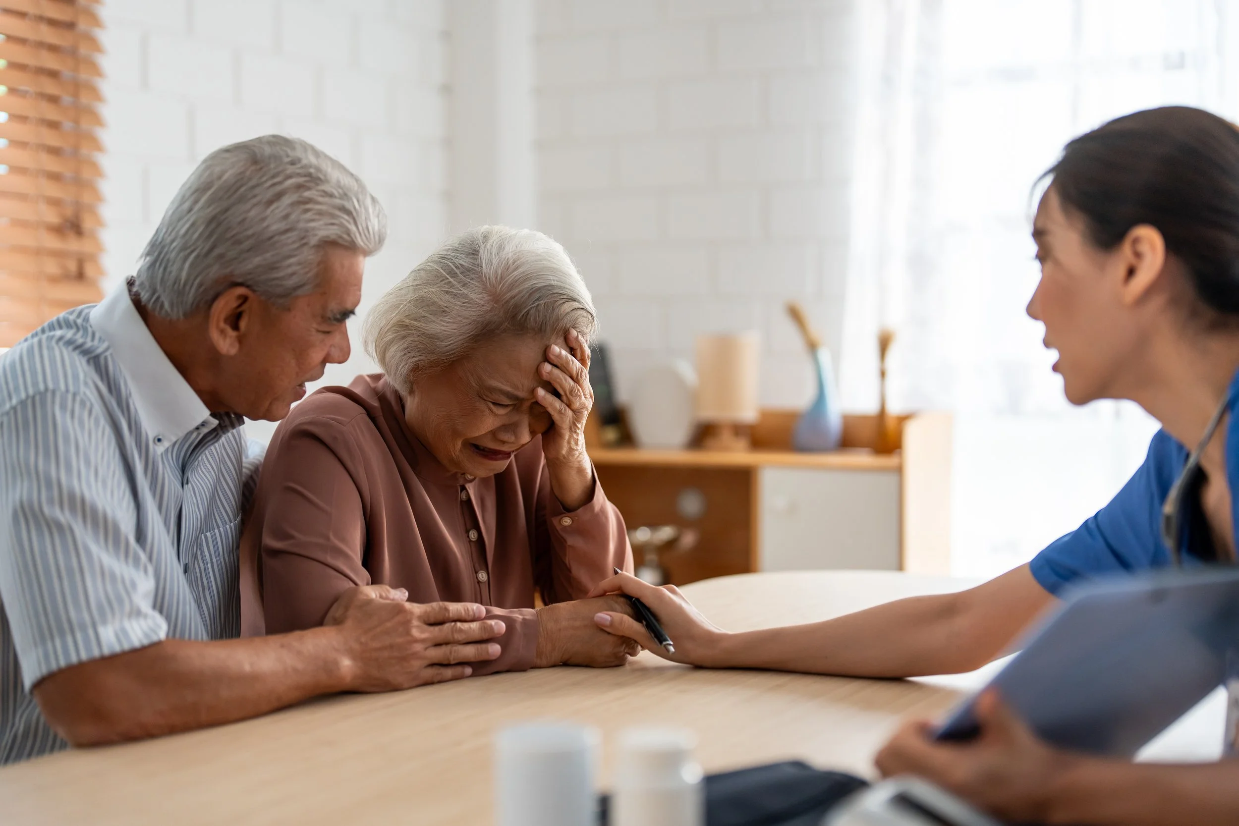 An elderly woman is crying and holding her forehead while being comforted by an elderly man and a healthcare professional during a consultation.
