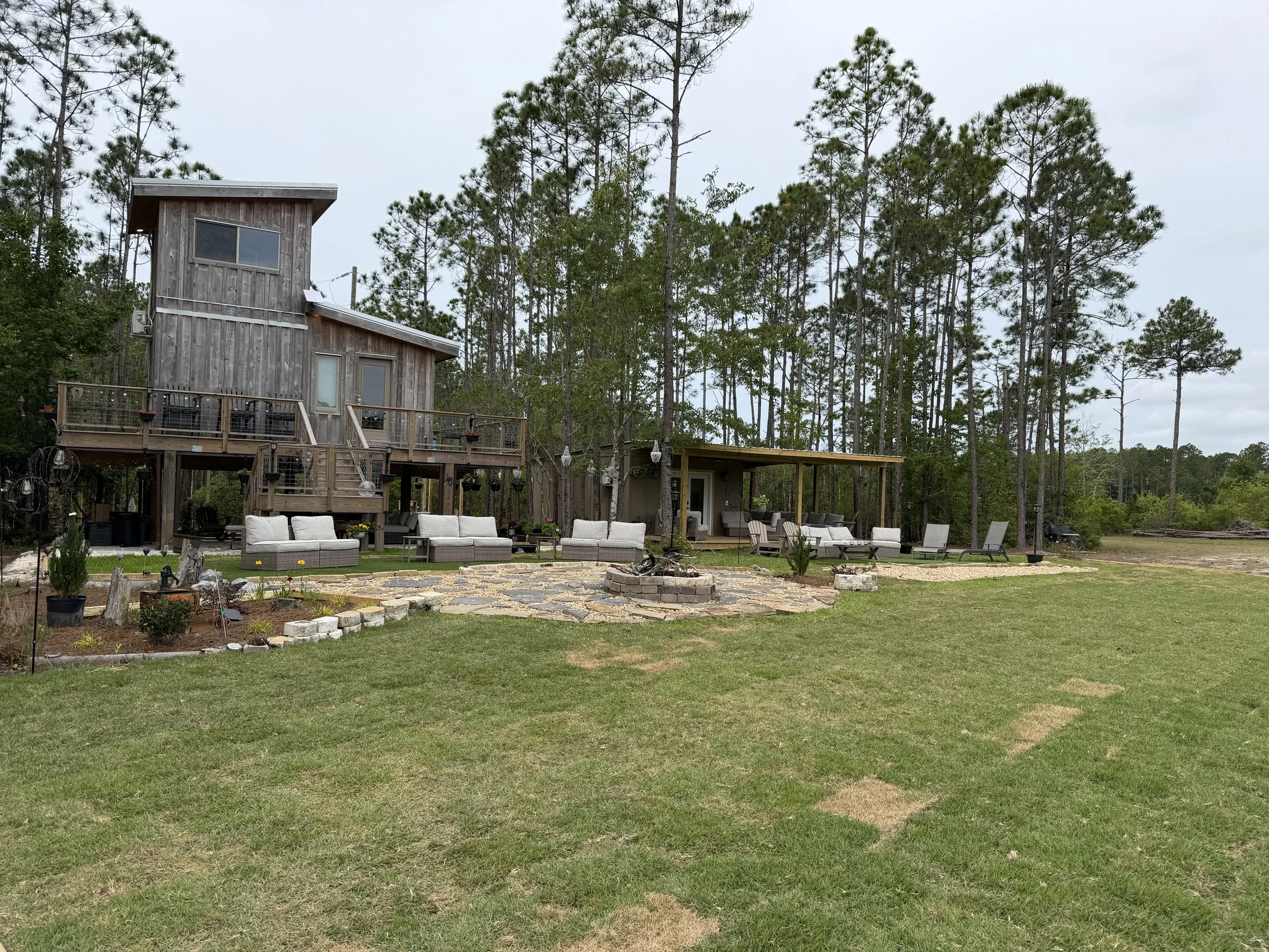 A backyard with a wooden house on stilts, surrounded by tall pine trees, outdoor furniture, and a landscaped garden area with a curved stone pathway.