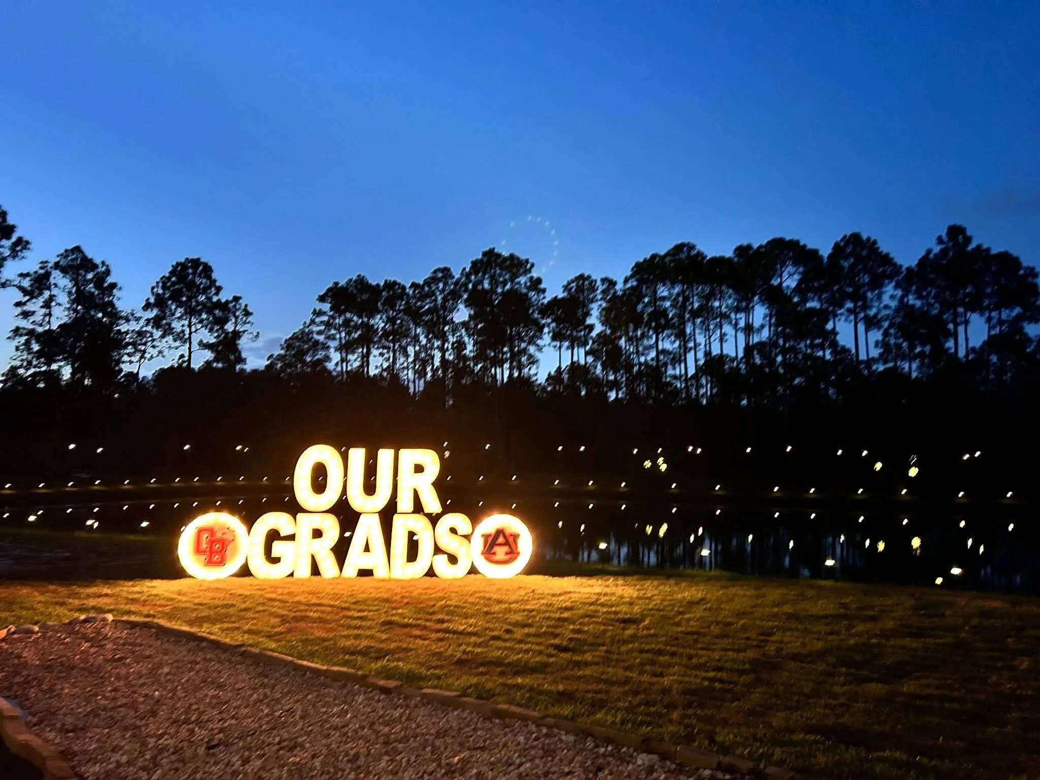 Decorative sign saying "Our Graduates" with graduation caps at night, illuminated lights, and trees in the background.