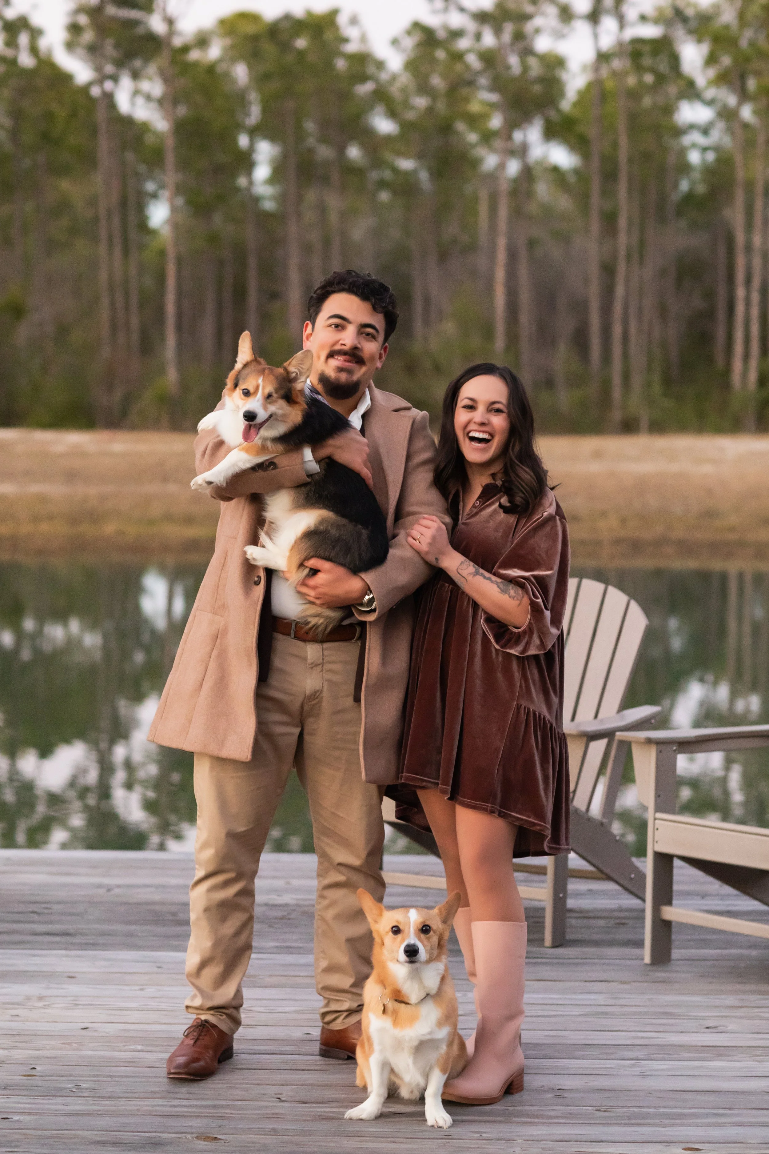 A man and woman standing on a wooden dock outdoors, smiling, with two dogs, one of which is sitting on the dock, and the other is being held by the man. The background features a calm lake and a forest of tall trees.