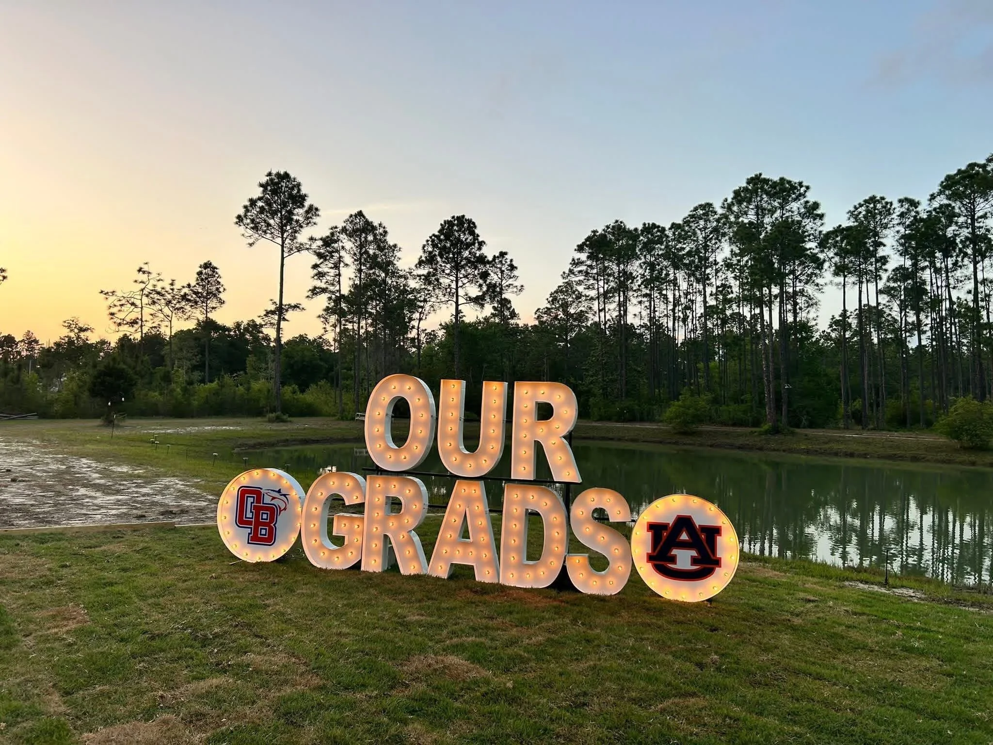 Large decorative sign reading 'OUR GRADS' with university logos, set outdoors near a water body with tall trees in the background at dusk.