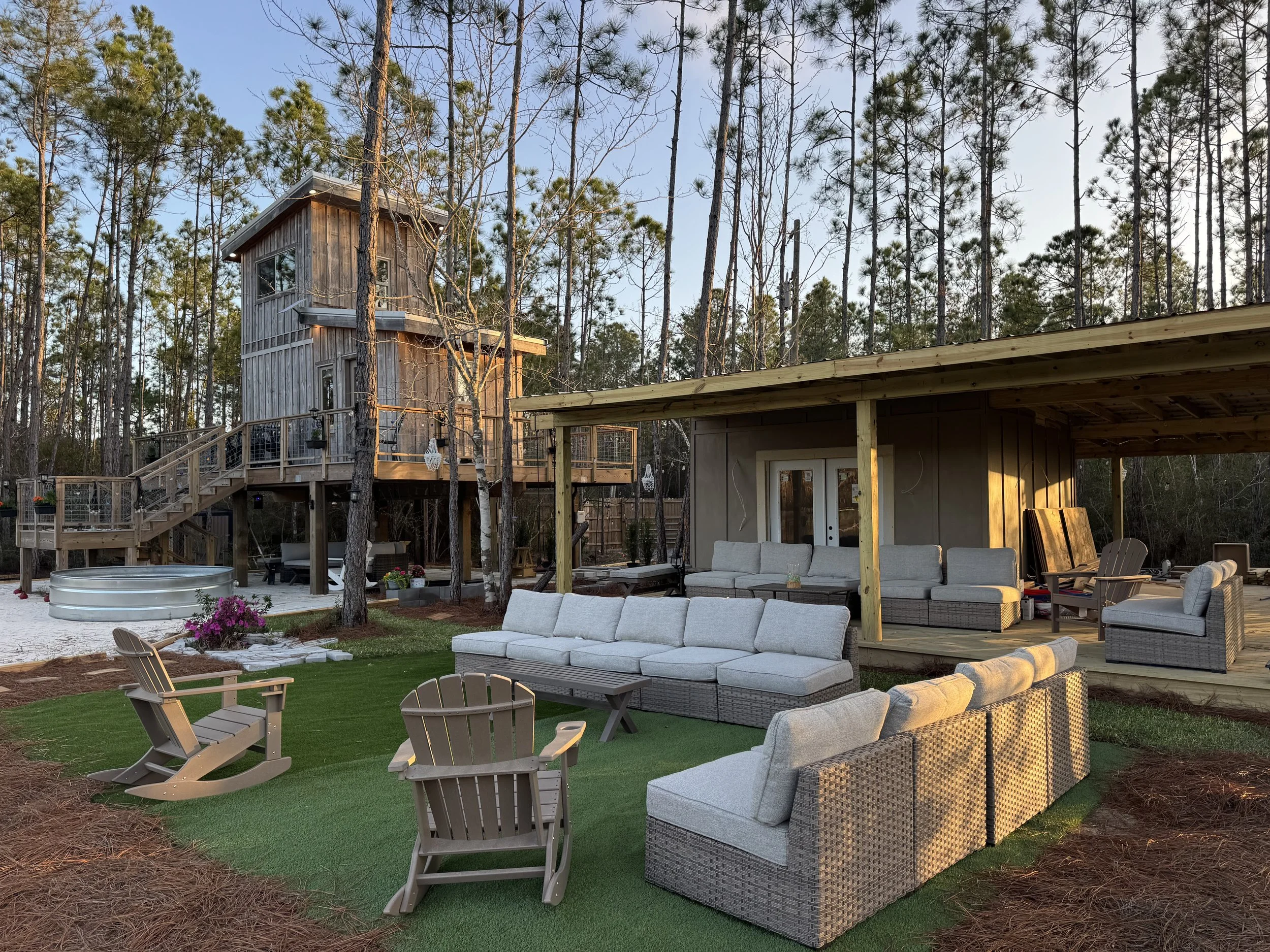 An outdoor patio area with modern furniture, including lounge chairs and couches, under a wooden pergola, surrounded by pine trees, with a two-story wooden house in the background.
