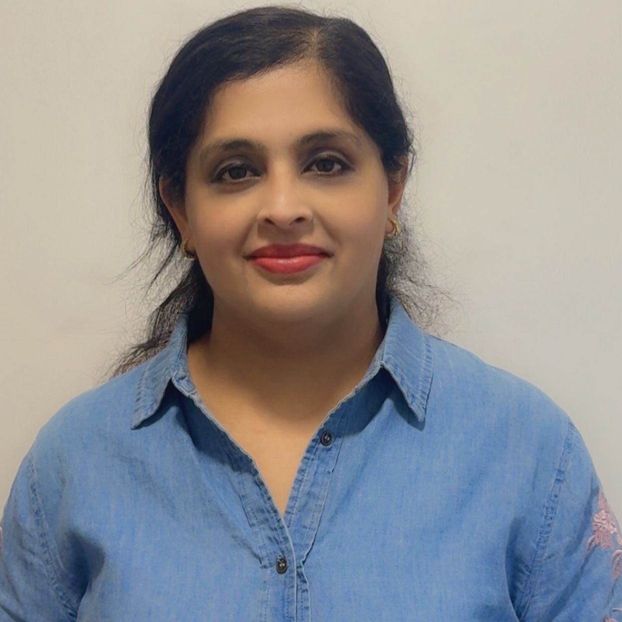 Portrait of a woman with dark hair, wearing a blue denim shirt, smiling, against a plain light-colored background.