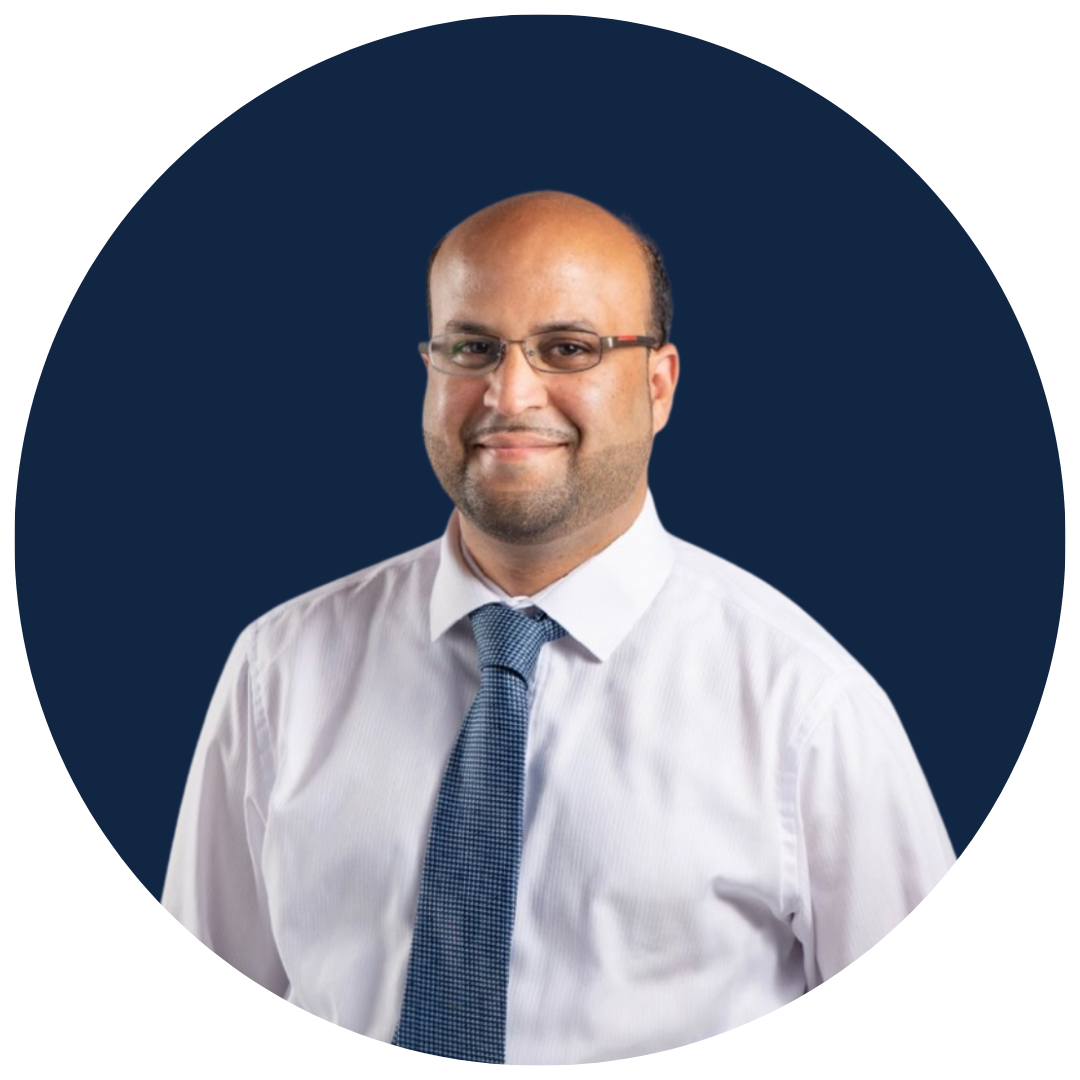 A man smiling wearing glasses, a white dress shirt, and a blue tie, posing against a dark blue background.