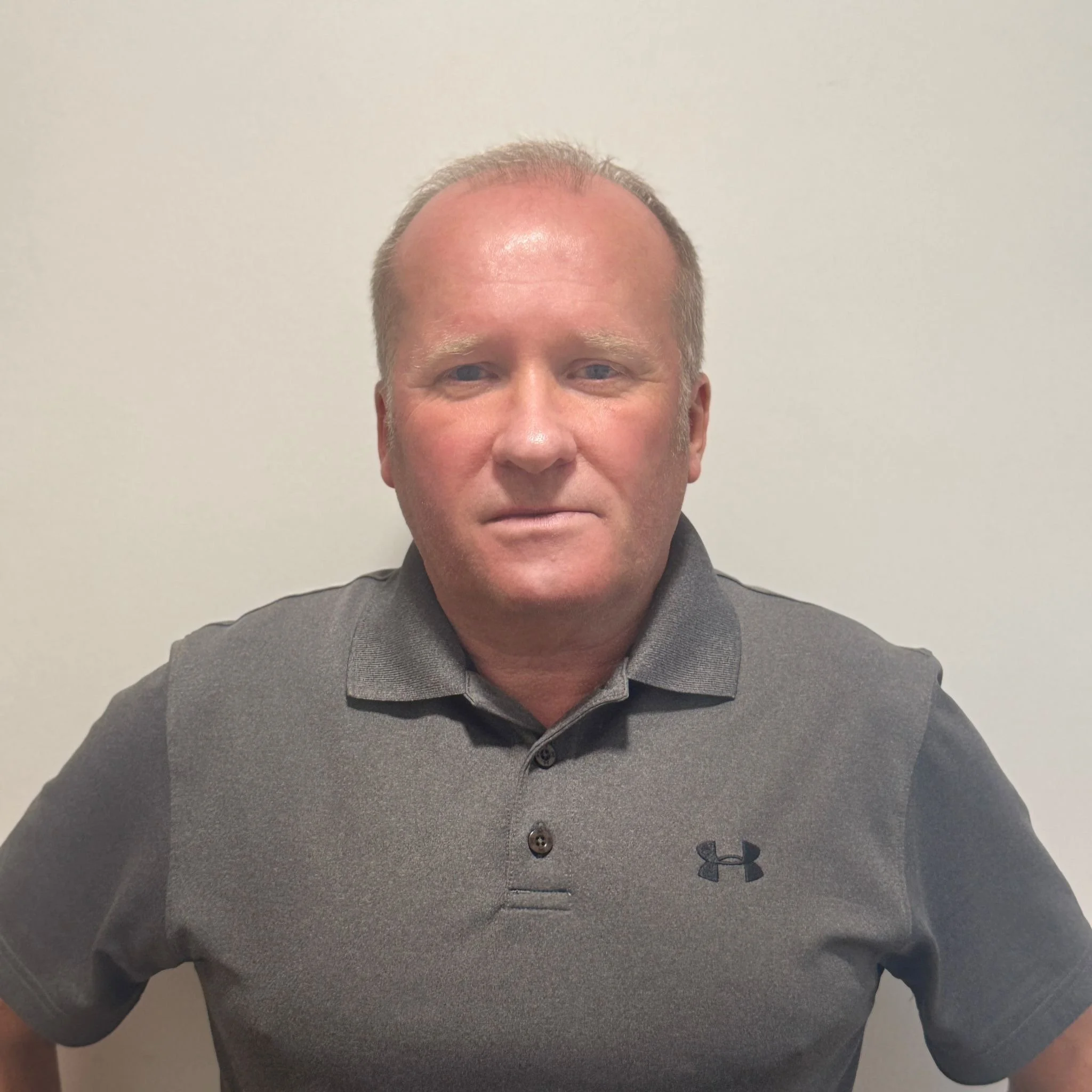 A middle-aged man with short, light-colored hair wearing a gray Under Armour polo shirt standing against a plain white wall.