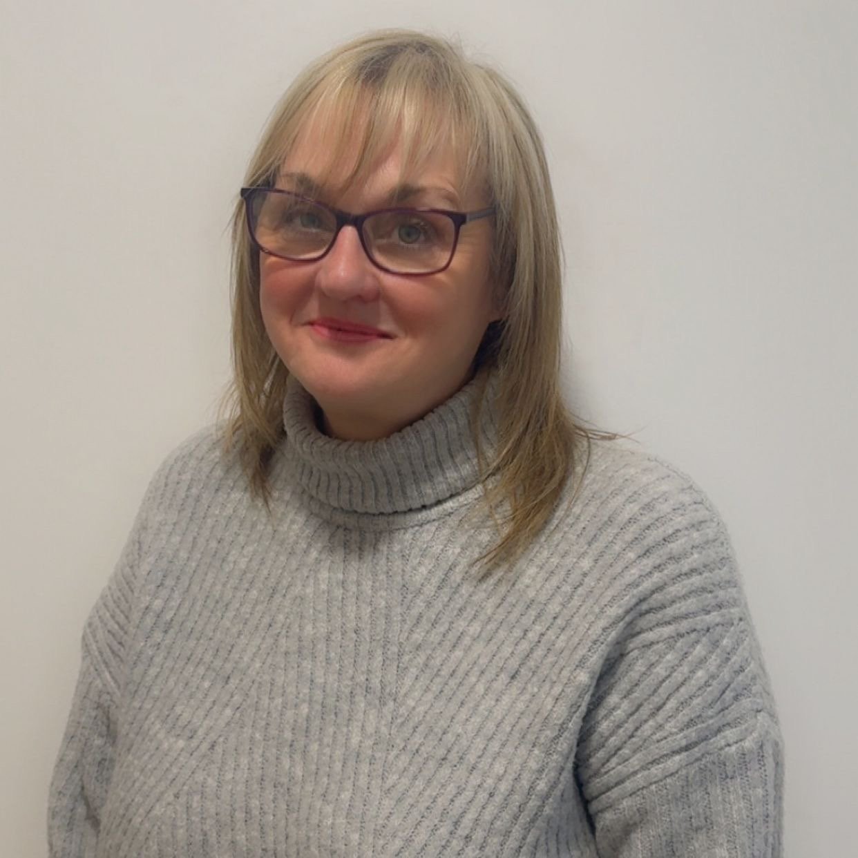 A woman with blonde hair and glasses wearing a gray turtleneck sweater, standing against a white wall.