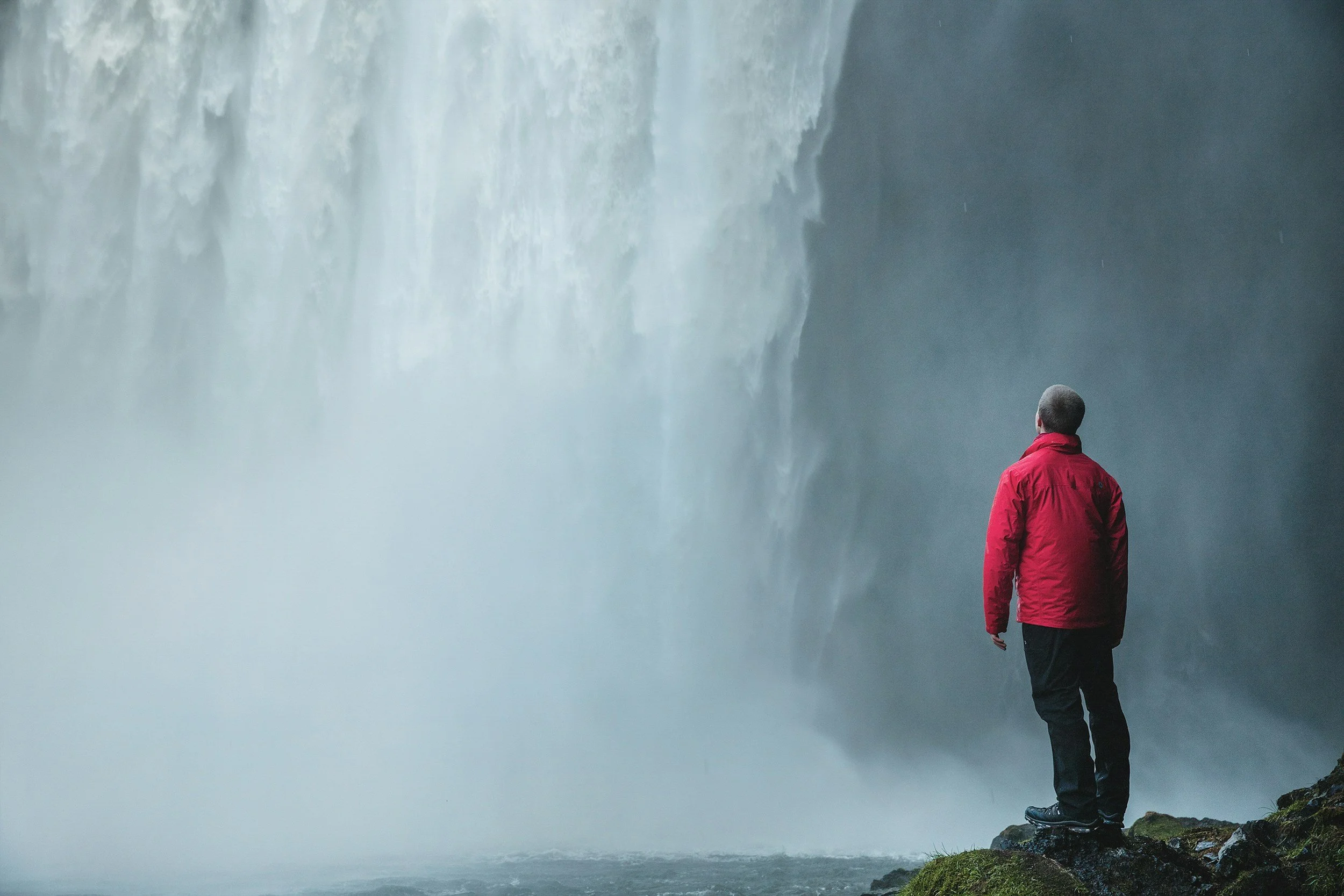 Man standing on rocks in front of a large waterfall wearing a red jacket and black pants.
