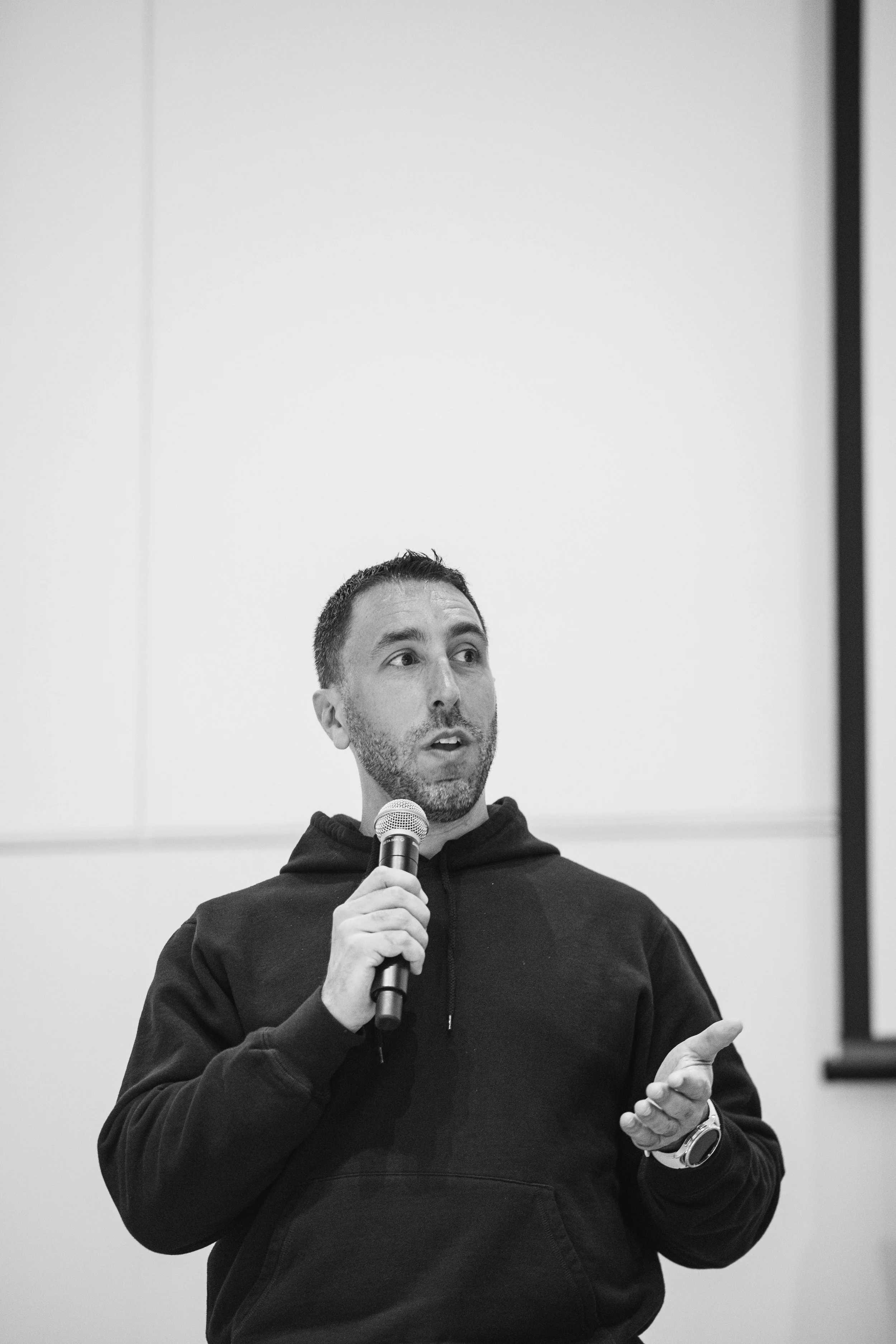 A man speaking into a microphone during a presentation or speech, wearing a black hoodie and watch, with a plain wall and partially visible screen behind him.
