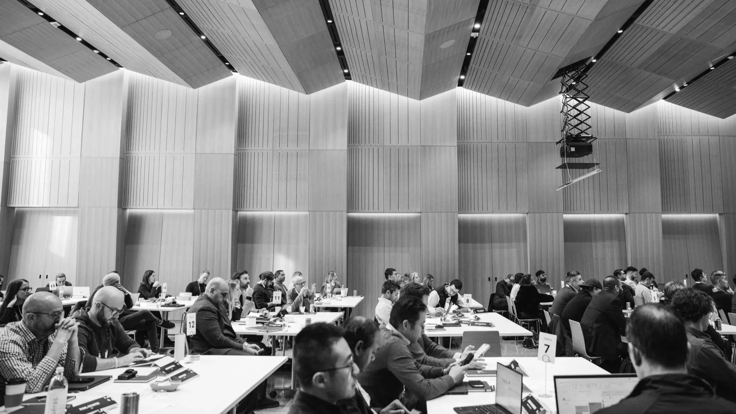 A large conference room filled with attendees sitting at long tables, some using laptops and others looking at their phones. The room has a high ceiling with wooden panels and vertical wall panels, with lighting strip accents and mounted equipment hanging from the ceiling.