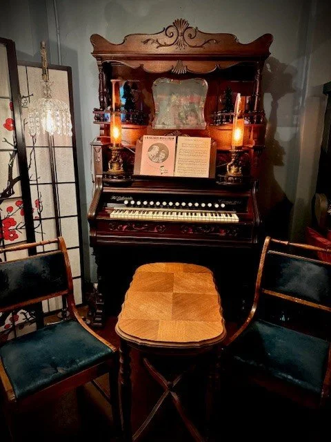 Vintage upright piano with an open sheet music book, flanked by two candles in holders, with two chairs on either side.