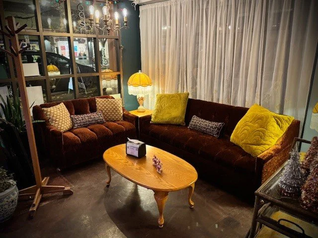 A cozy living room with two vintage-style velvet sofas, one in dark red and the other in dark brown, decorated with colorful throw pillows. A wooden coffee table with curved legs sits in front of the sofas, with a small decorative item and a purple flower on it. A yellow table lamp with a white shade provides warm lighting. Behind the sofas, there's a large window with sheer white curtains, and a glass partition that separates the living room from an entryway or patio.