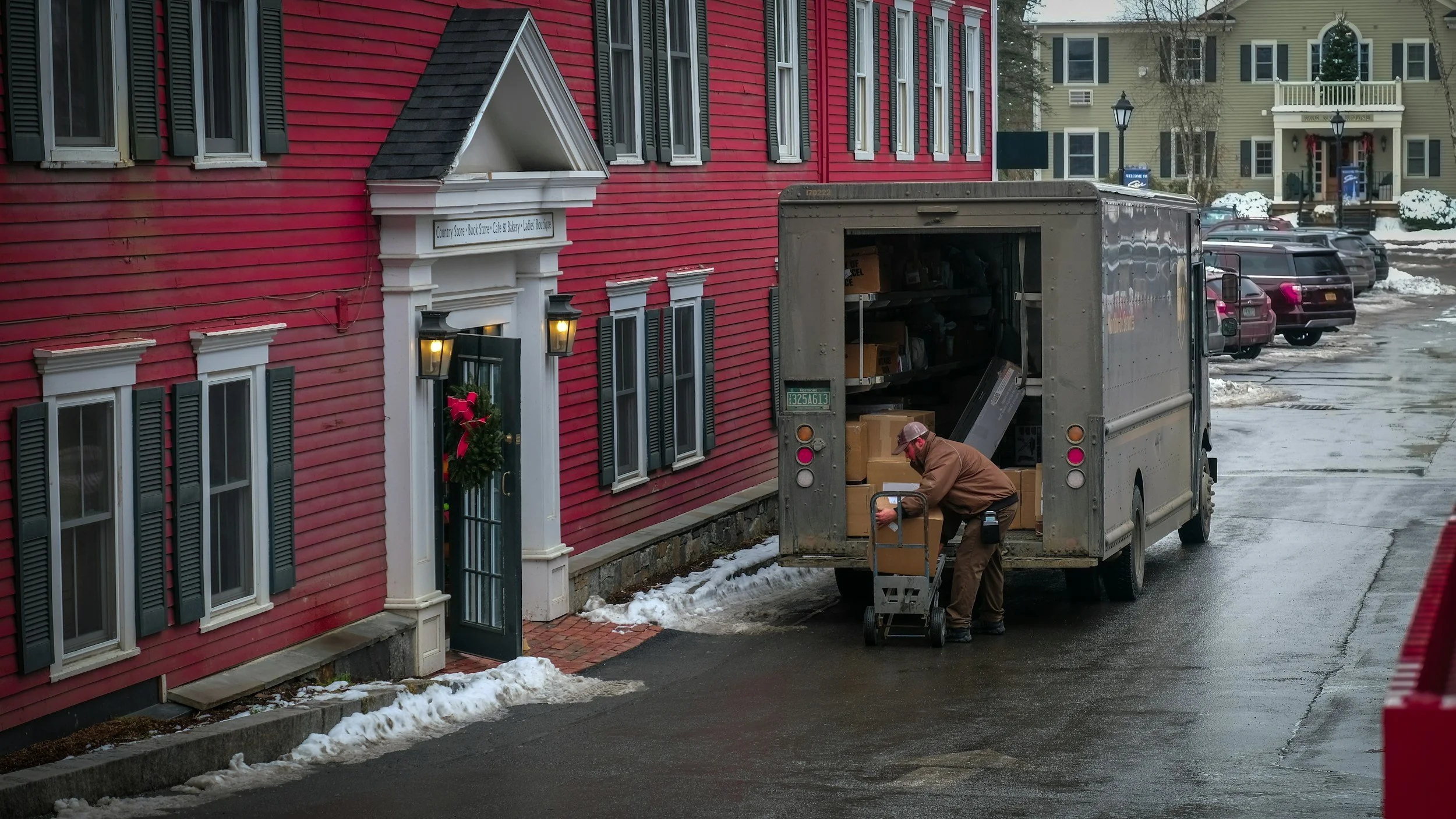 A man loading boxes into a delivery truck outside a red building with green shutters and wreaths on the door, on a snowy day.