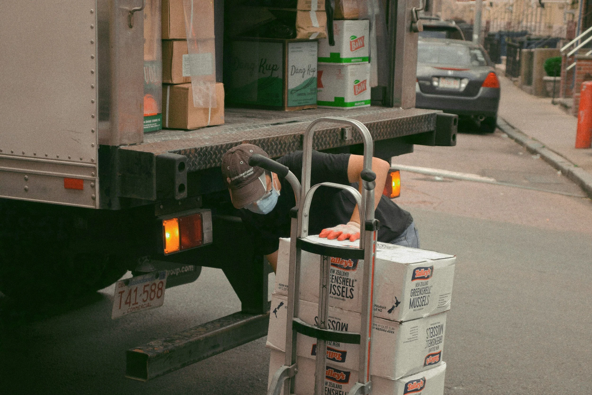A worker wearing a face mask and black shirt is loading boxes of greenshell mussels onto a delivery truck, using a small hand truck on a city street.