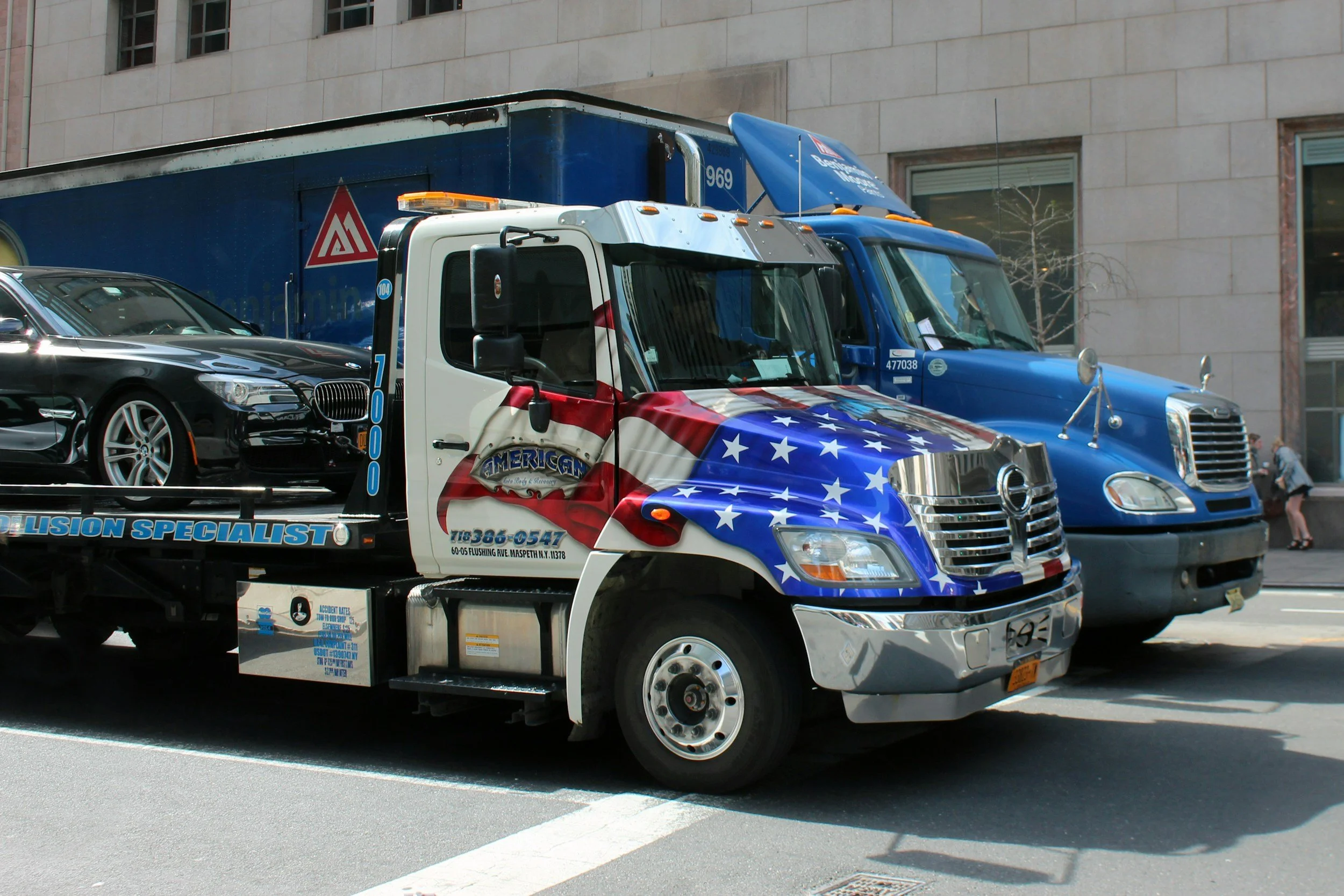 A tow truck decorated with an American flag design parked on the street alongside a blue truck and a black car.