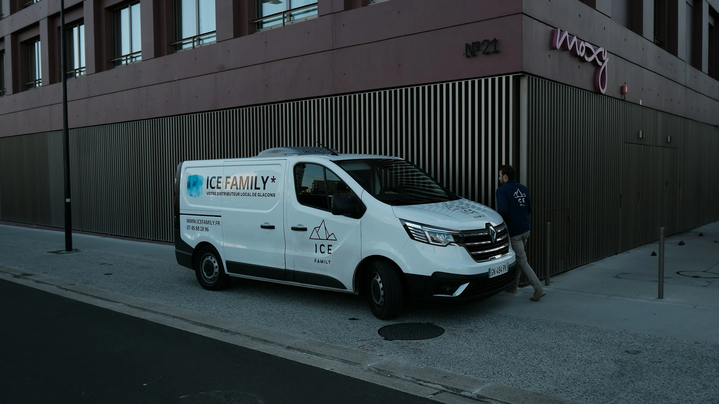 A white ICE Family delivery van parked on a city sidewalk next to a modern building with the Macy's sign visible. A person wearing a blue jacket is standing near the van.