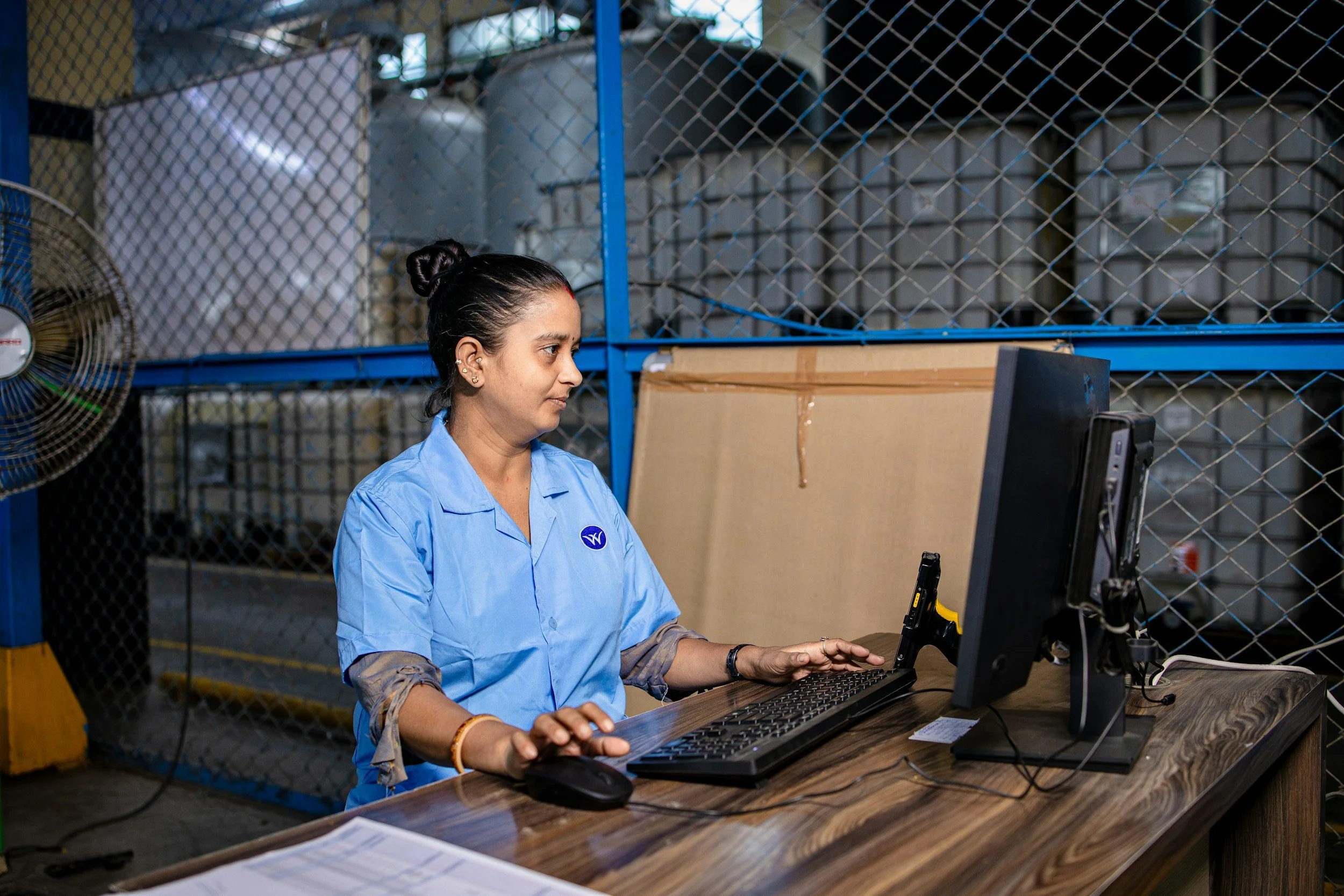 Woman in blue work uniform sitting at a desk using a computer in a warehouse or industrial setting.