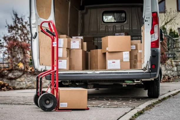 Open delivery van filled with boxes and a red hand truck on the sidewalk.