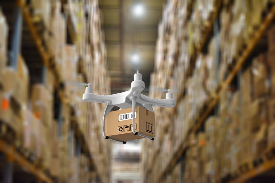 A drone carrying a small cardboard box through a warehouse aisle with tall shelves filled with boxes.