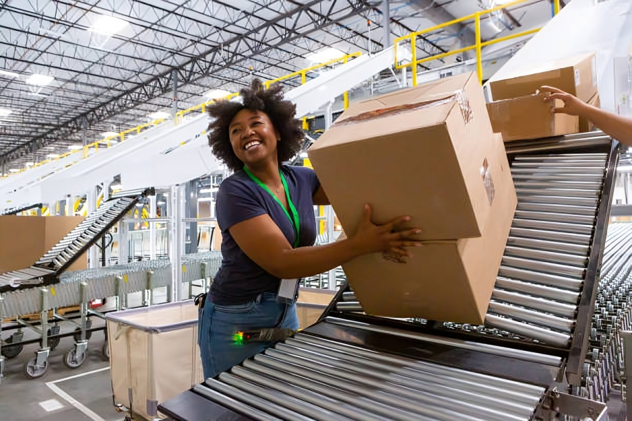 A woman working in a warehouse, loading a large cardboard box onto a conveyor belt, with other boxes and warehouse equipment around her.