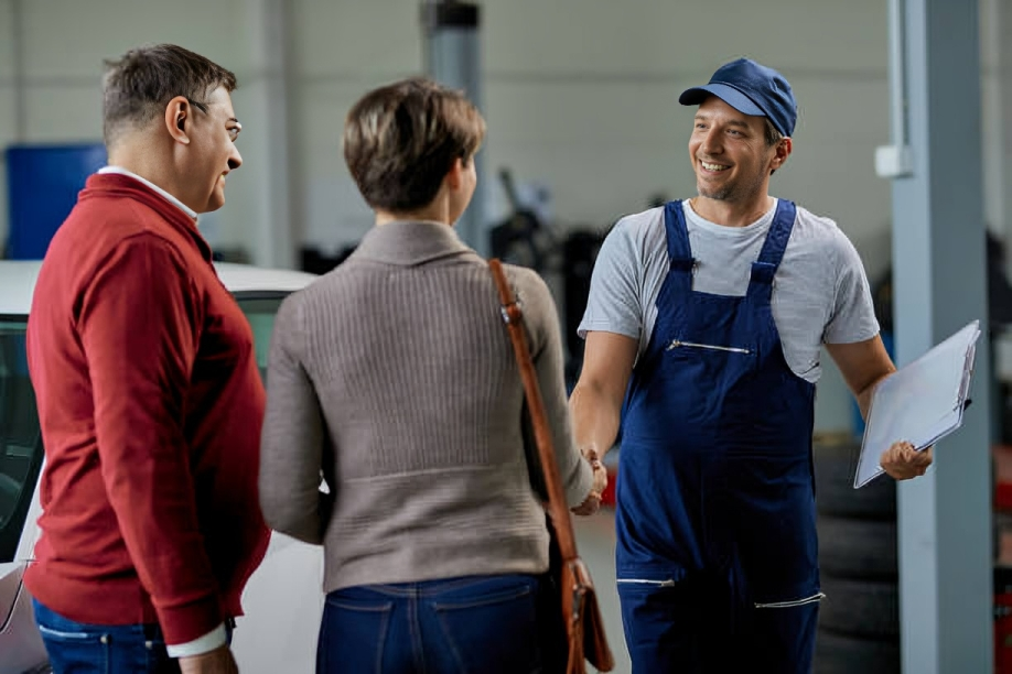 A man in a blue work uniform and cap shaking hands with two women in a garage or automotive repair shop.