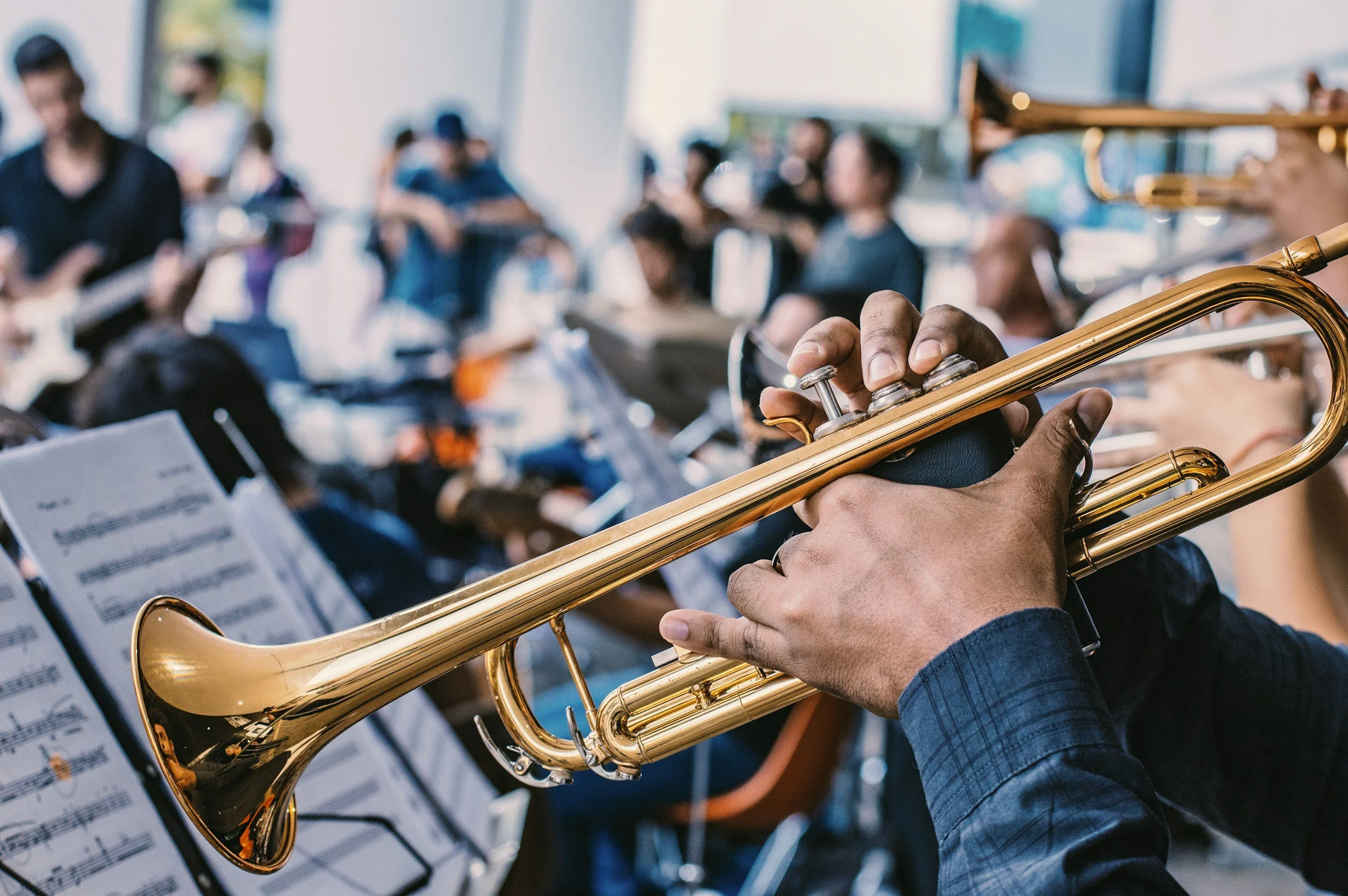 Close-up of a musician's hand playing a brass trumpet during an indoor orchestra concert, with other band members and music stands visible in the blurred background.