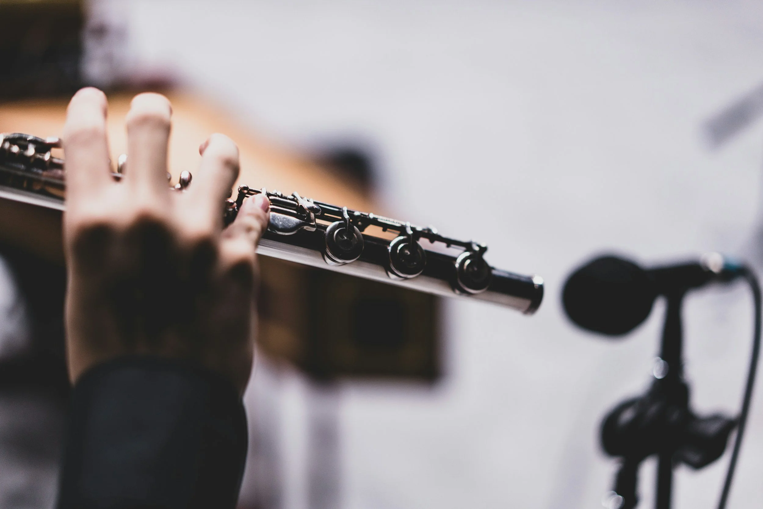 Close-up of a person's hand playing a black and silver flute, with a microphone on a stand in the background.