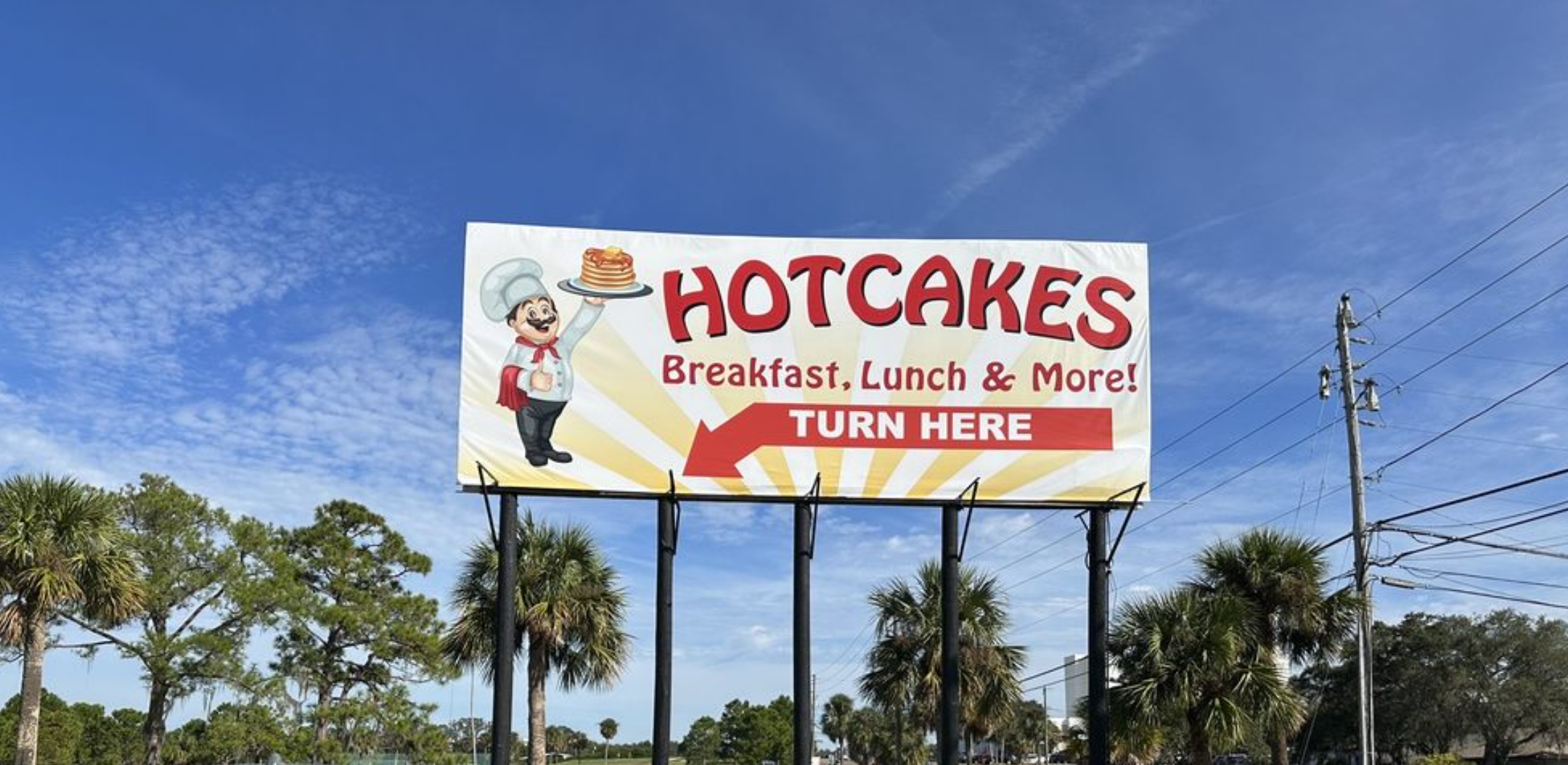 Billboard advertising a restaurant called Hotcakes with breakfast, lunch, and more. Features a cartoon chef holding a plate with a stack of pancakes. An arrow points left with the text 'Turn Here'.