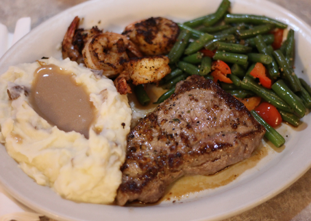 A plate of food with mashed potatoes topped with gravy, grilled shrimp, green beans with red peppers, and a grilled steak.