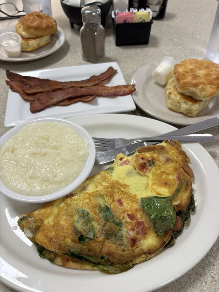 Plate with omelet containing spinach and tomatoes, mashed potatoes, a biscuit with butter, and a side of bacon.