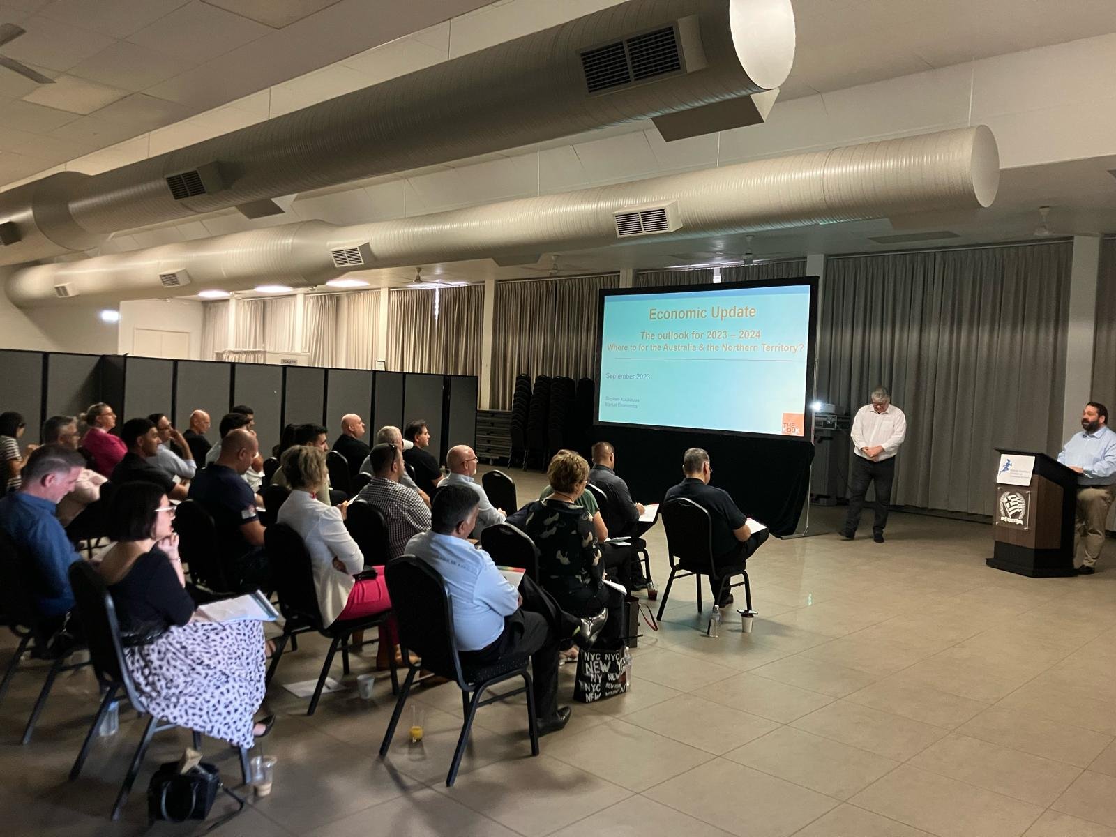 A group of people attending a presentation in a conference room. The presentation slide on the screen reads 'Economic Update' and mentions the outlook for 2023-2024.