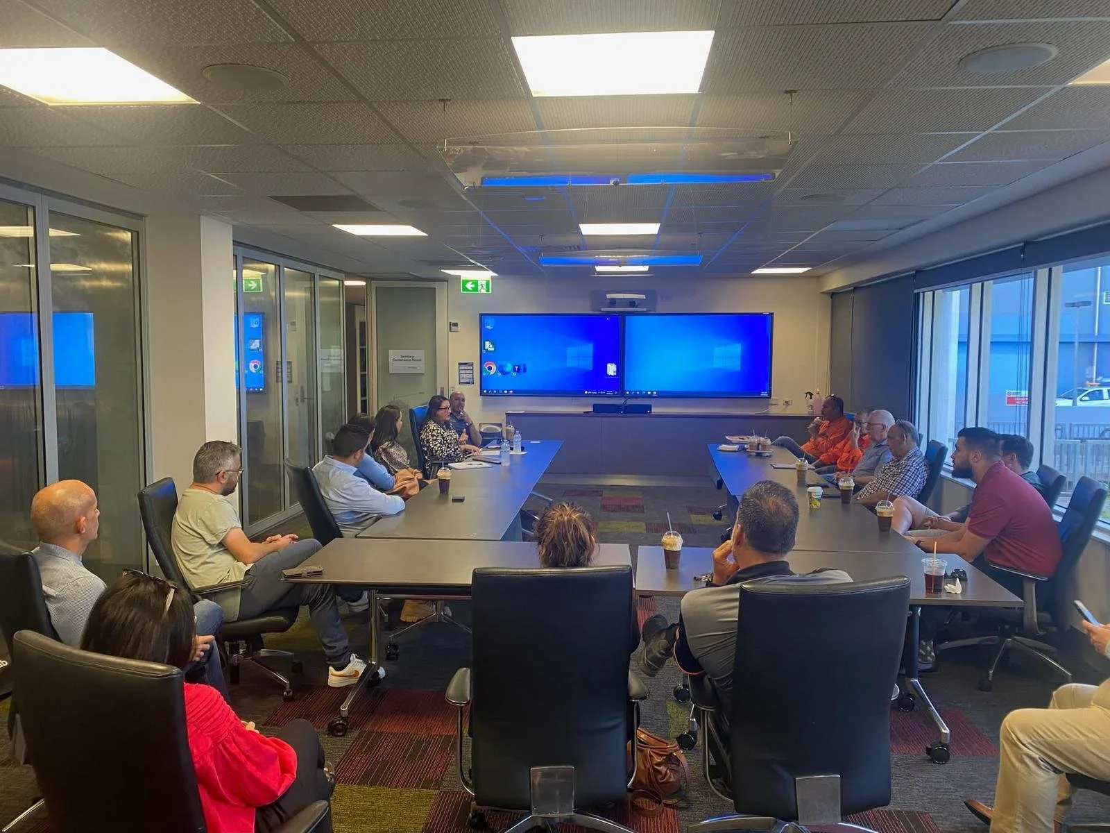 A group of people attending a meeting or presentation in a modern conference room with large windows, a long table, and multiple screens displaying the Windows desktop.