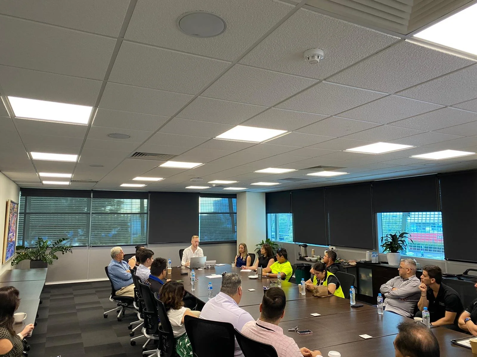 A business meeting in a conference room with multiple attendees sitting around a large table, some of them taking notes or listening. A person is presenting at the head of the table. The room has large windows with blinds, potted plants, and artwork on the walls.