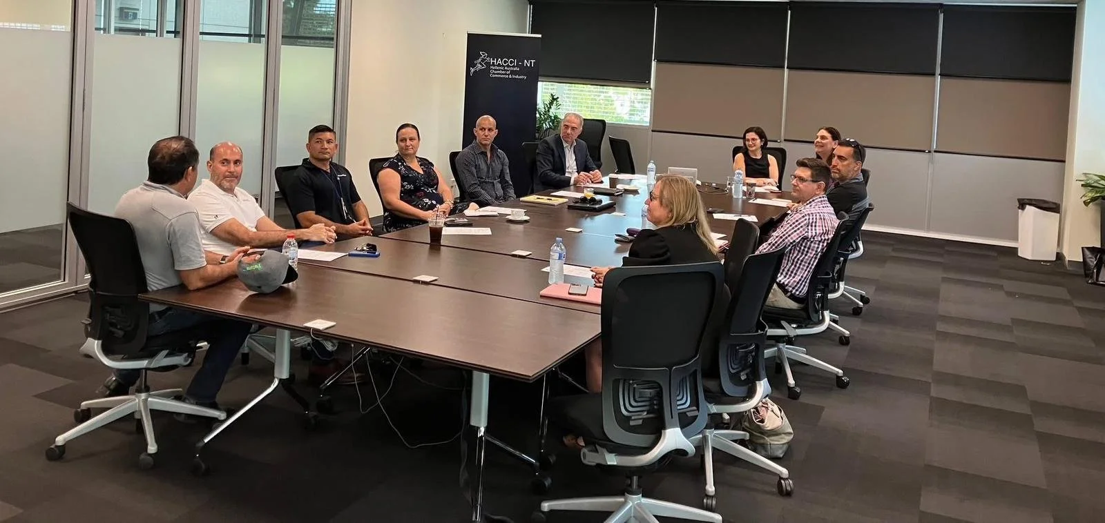 A group of twelve people attending a business meeting in a modern conference room with glass walls, table, chairs, and branded banner in the background.