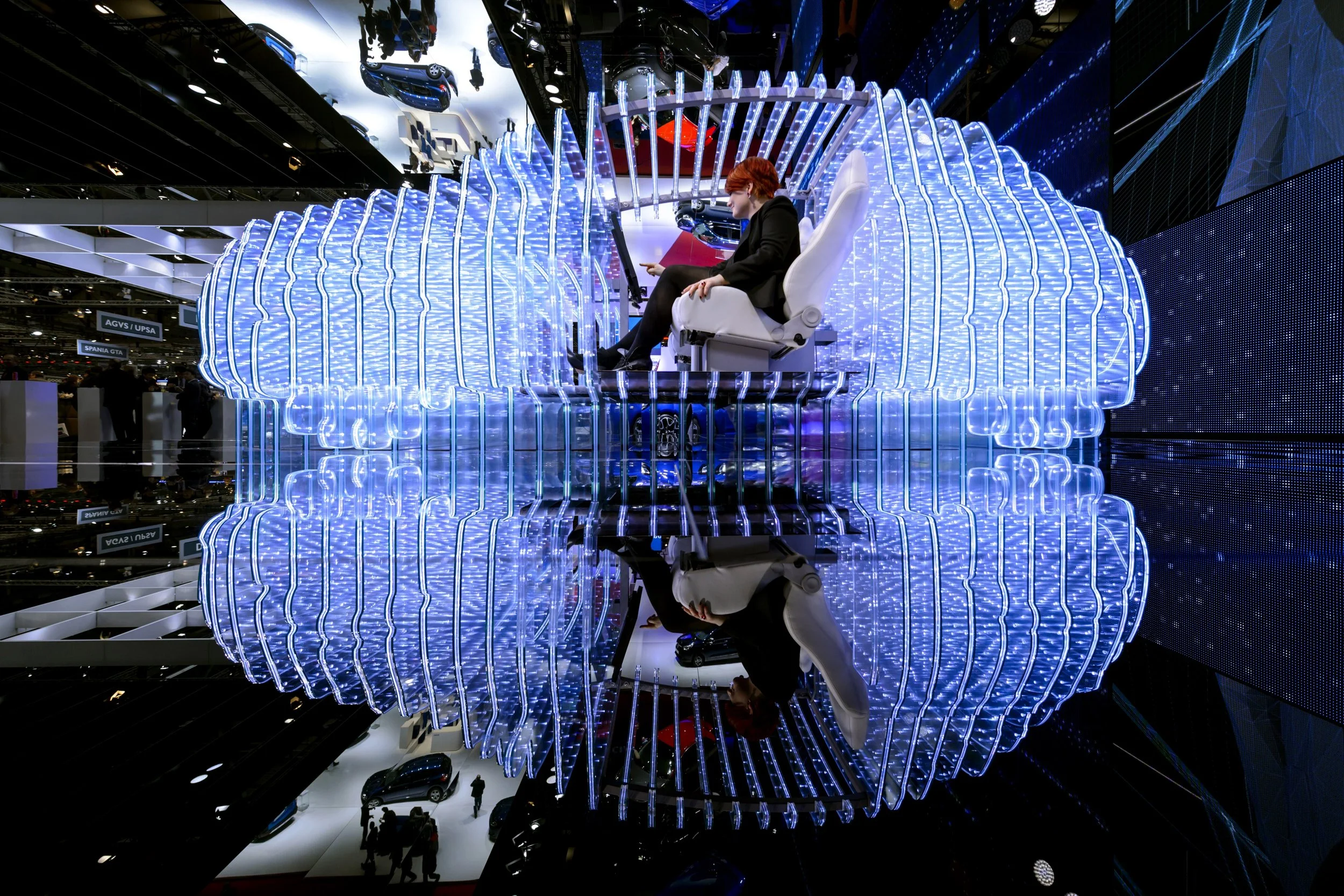 A person sitting in a futuristic chair surrounded by blue LED light structures, with their reflection visible on a shiny surface below, at an indoor automotive exhibition.