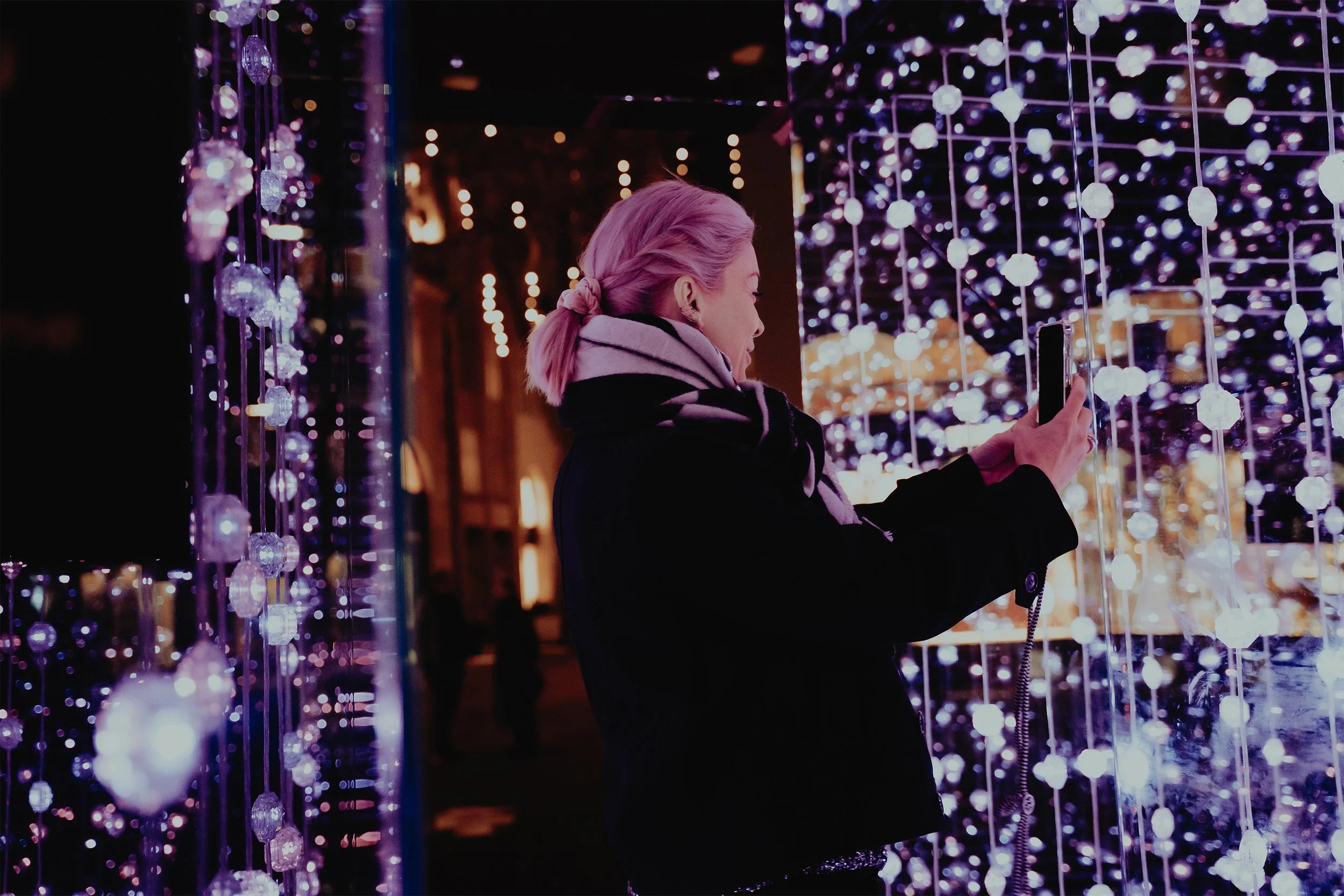 A woman with pink hair in a ponytail taking a photo with her phone among hanging purple and white lights at night.