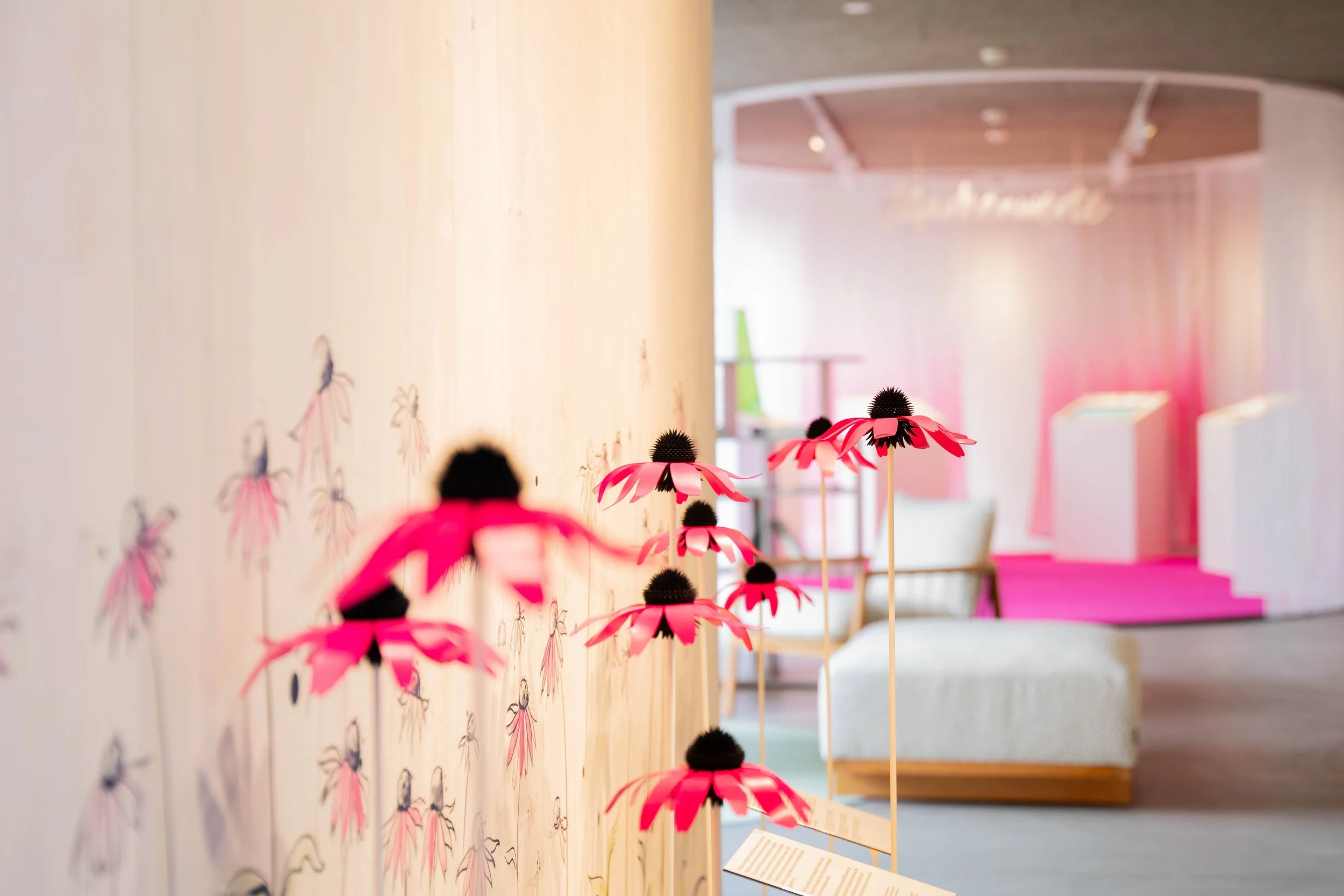 Exhibit of pink paper flowers with black centers on wooden sticks in a gallery, with pink and white decor in the background.