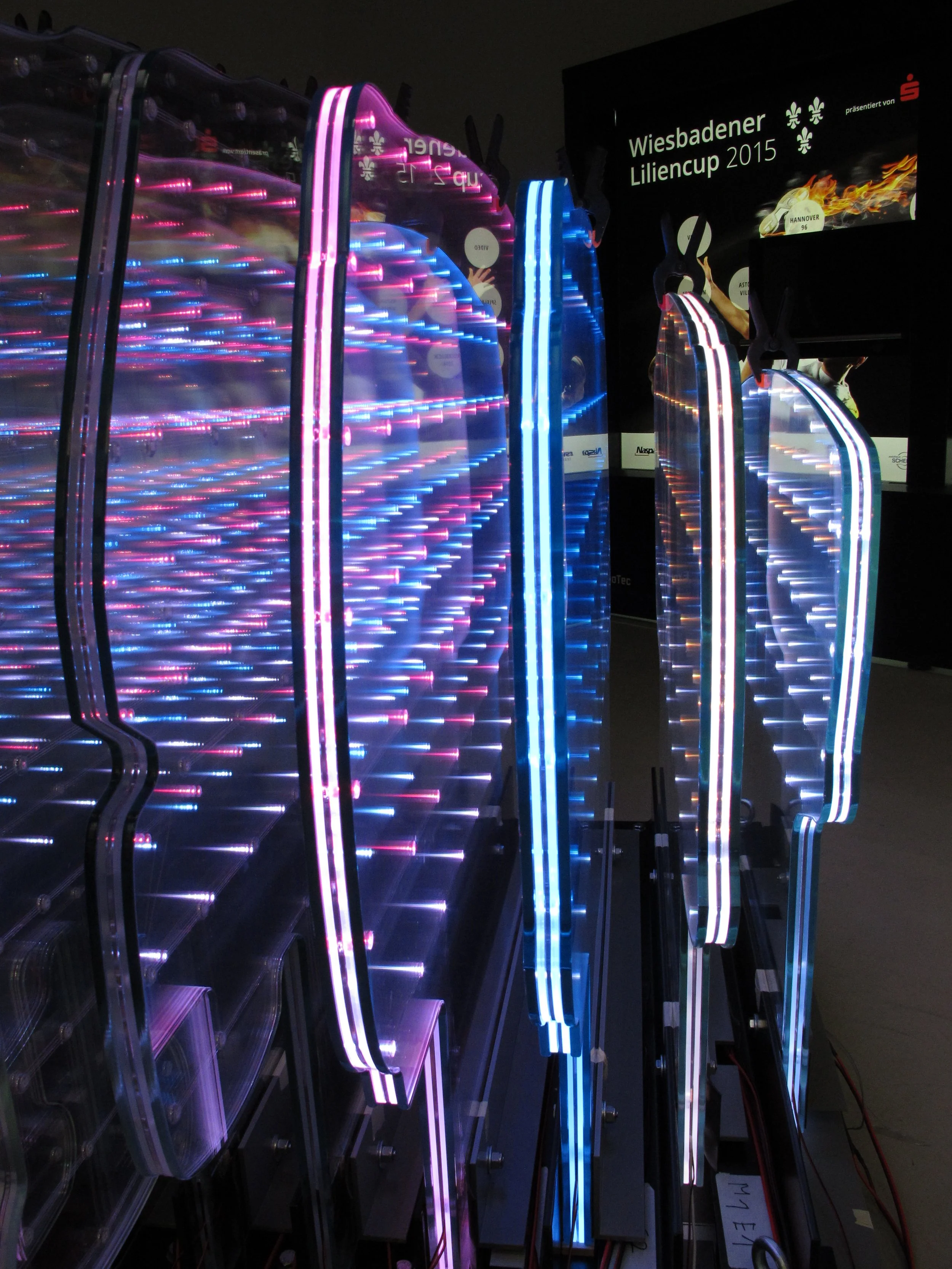 Close-up of colorful neon LED signs, some illuminated in pink, blue, and white, with a black background that includes a sign reading 'Wiesbadener Liliencup 2015'.