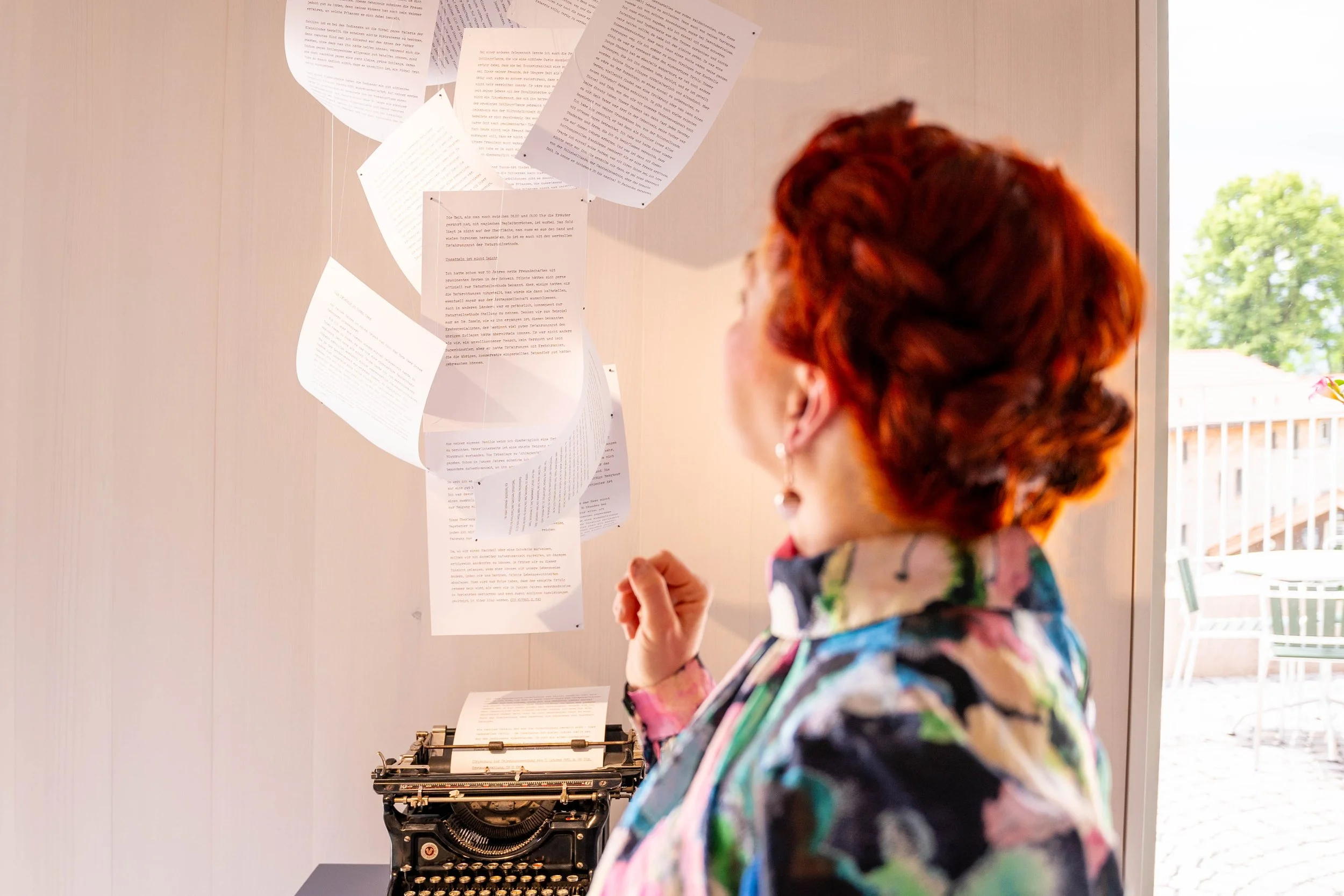 A woman with red hair, wearing a colorful patterned jacket, is looking at papers hanging on a wall and a typewriter on a table in front of her.
