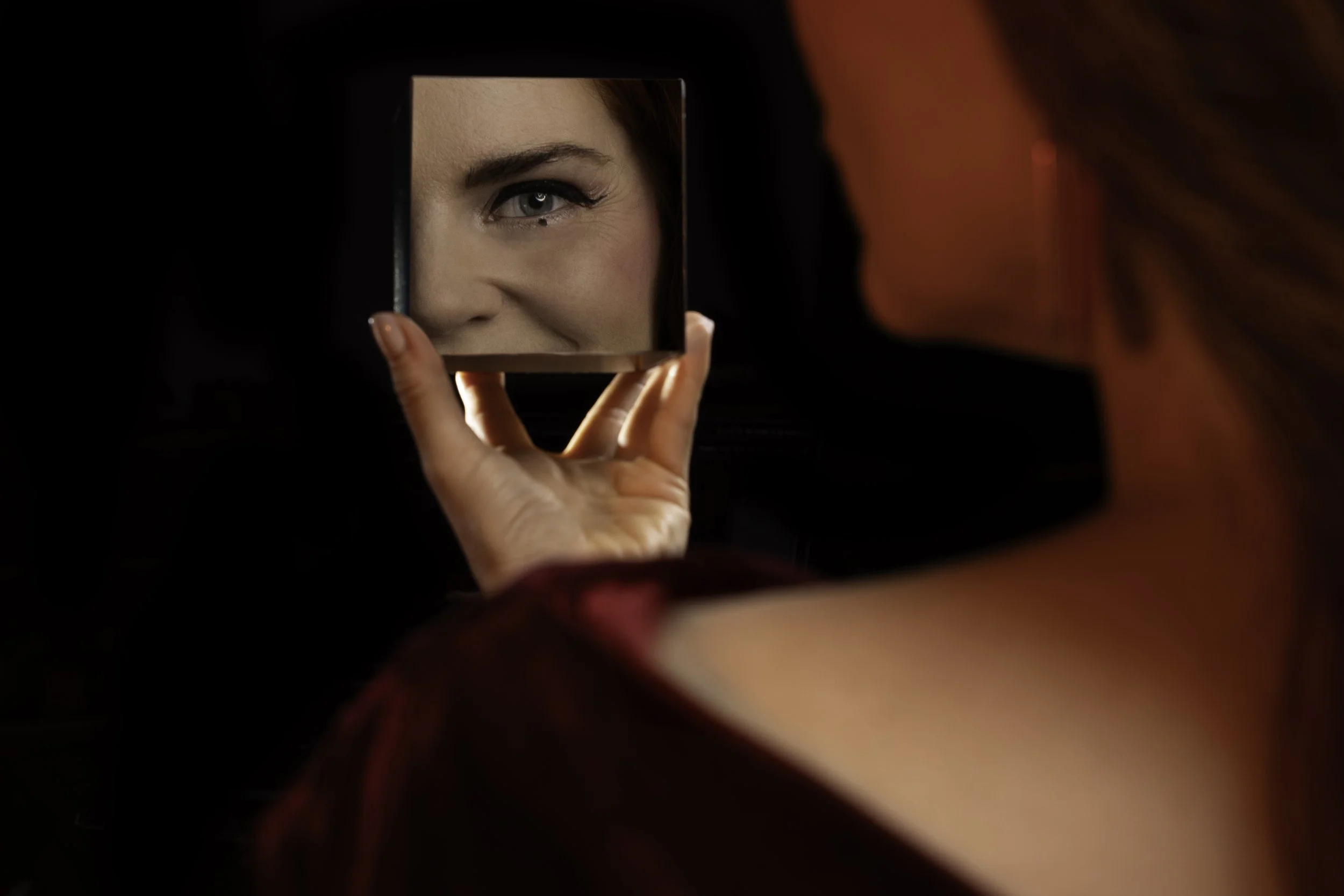 A woman with red hair holding a square mirror, reflecting her eye and part of her face, with the background dark.