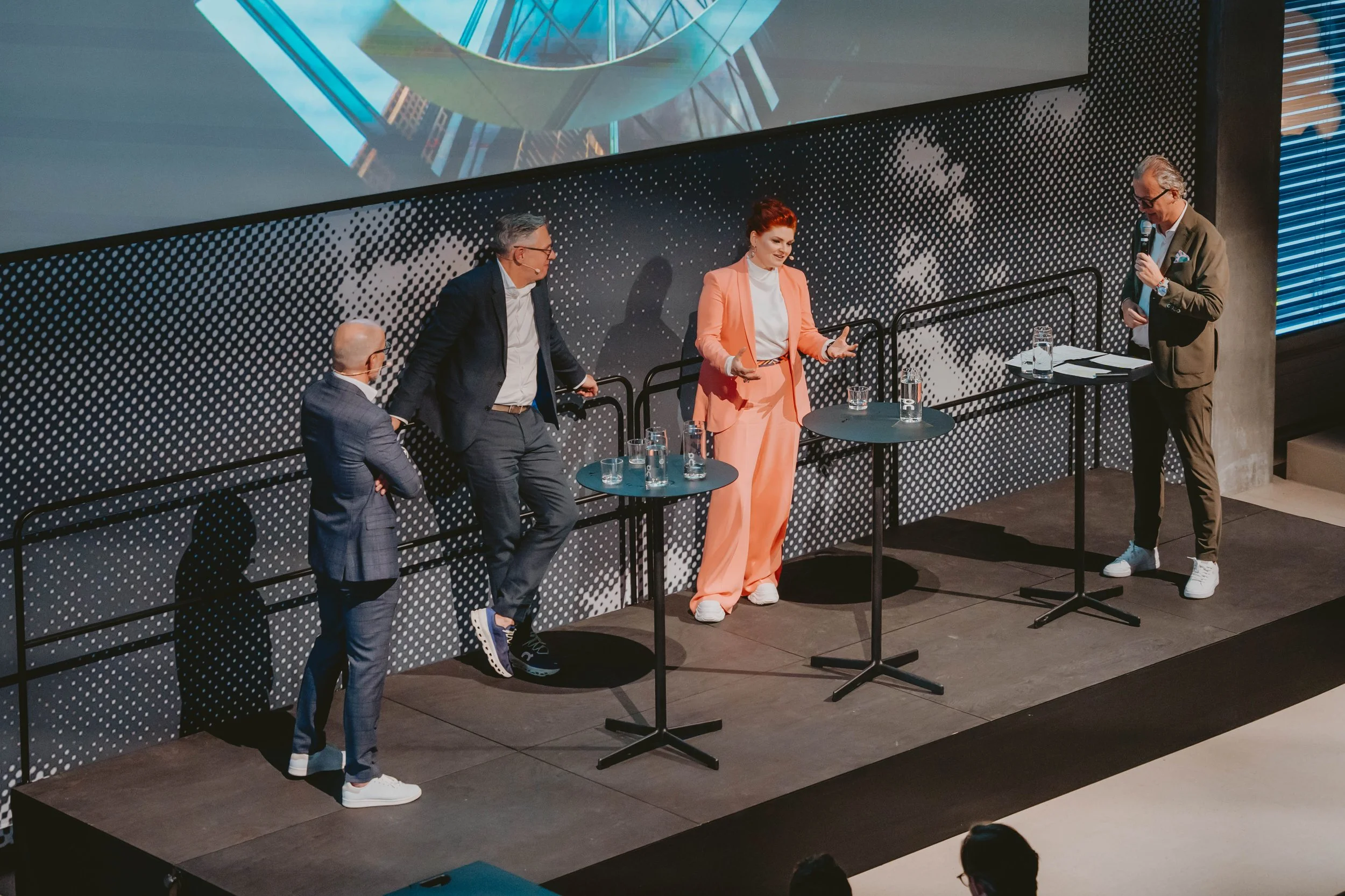 Four people on a stage in a panel discussion. Two men and one woman are standing, and one man is sitting. The woman is wearing a peach-colored suit, and the men are in suits. There are tables with glasses of water in front of them, and a large screen behind displaying a graphic.