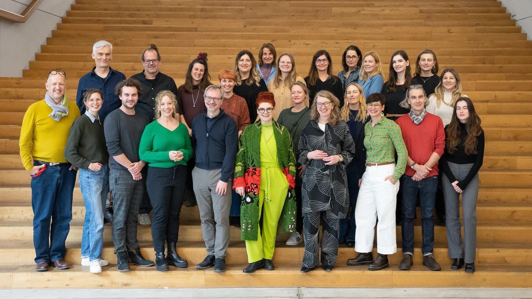 A group of diverse people standing on wooden stairs indoor, smiling at the camera.