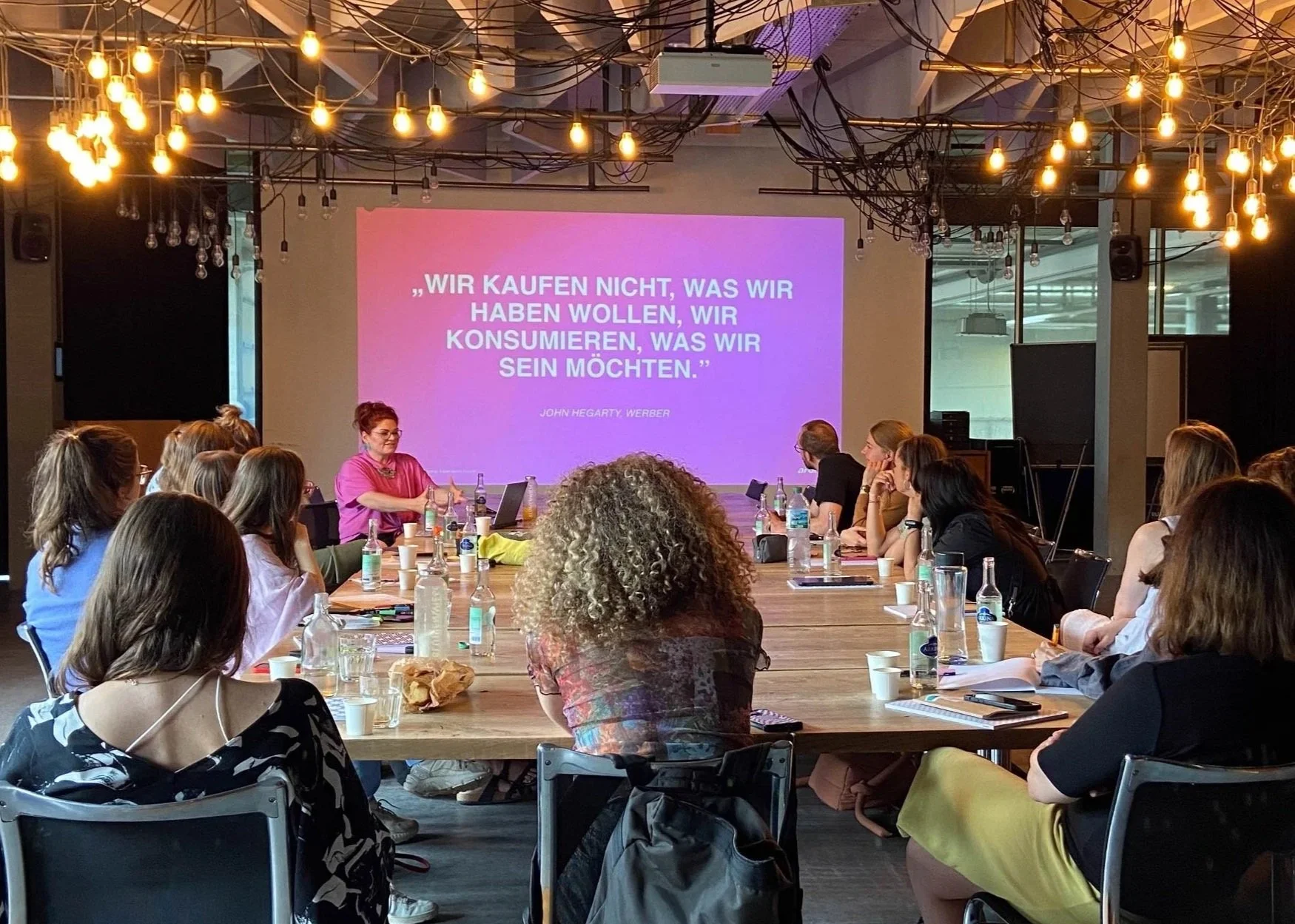 People attending a presentation or workshop in a modern conference room with a large screen displaying a quote in German, surrounded by hanging light bulbs.