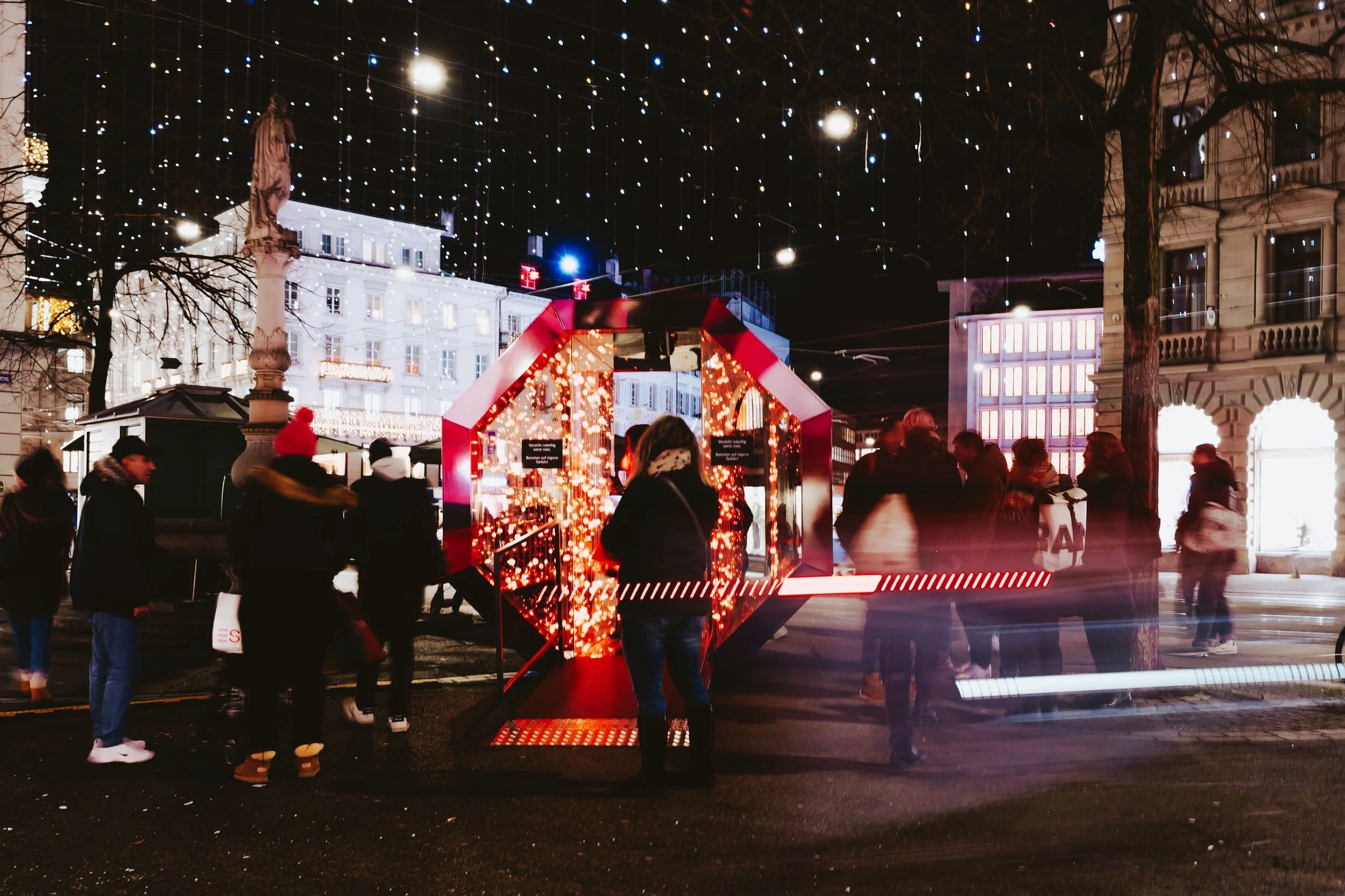 People walking and standing around a lit-up Christmas-themed display on a city street at night, with festive lights hanging overhead and historic buildings in the background.