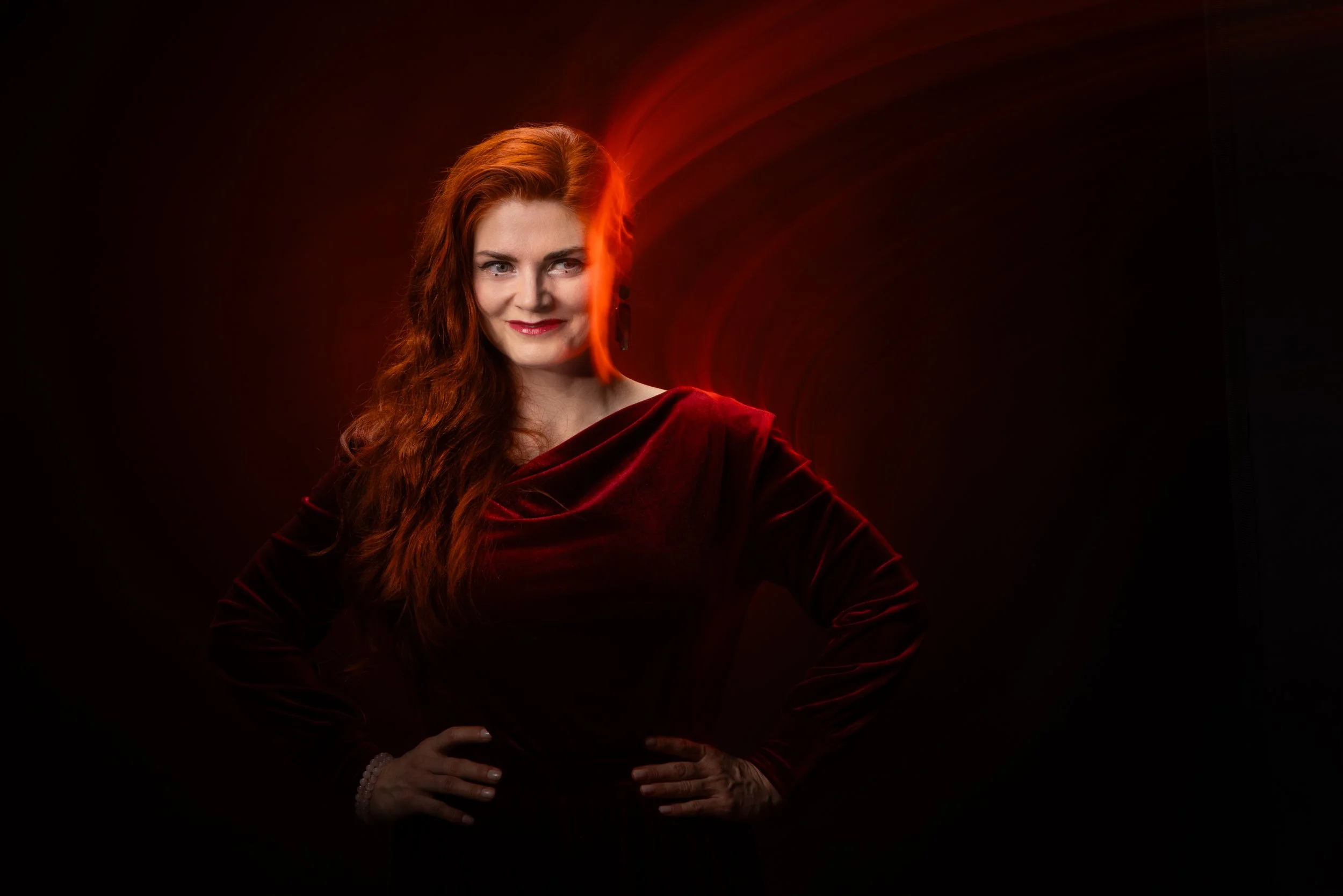 Red-haired woman in a dark red velvet dress with hands on hips, smiling confidently against a dark background with red light accents.