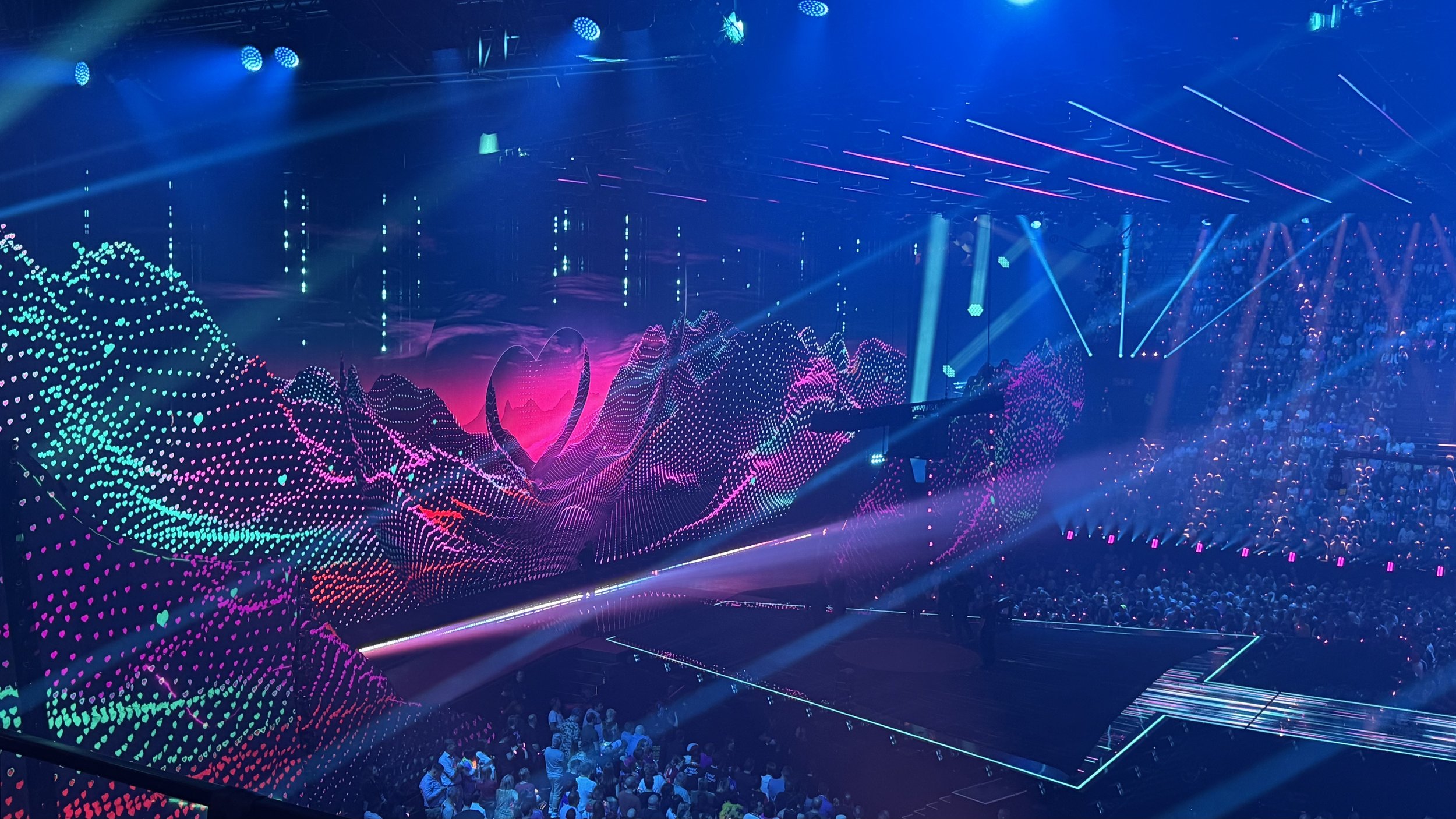 Concert stage with vibrant, colorful LED visual display of mountains and a heart in the background, with a large crowd watching.