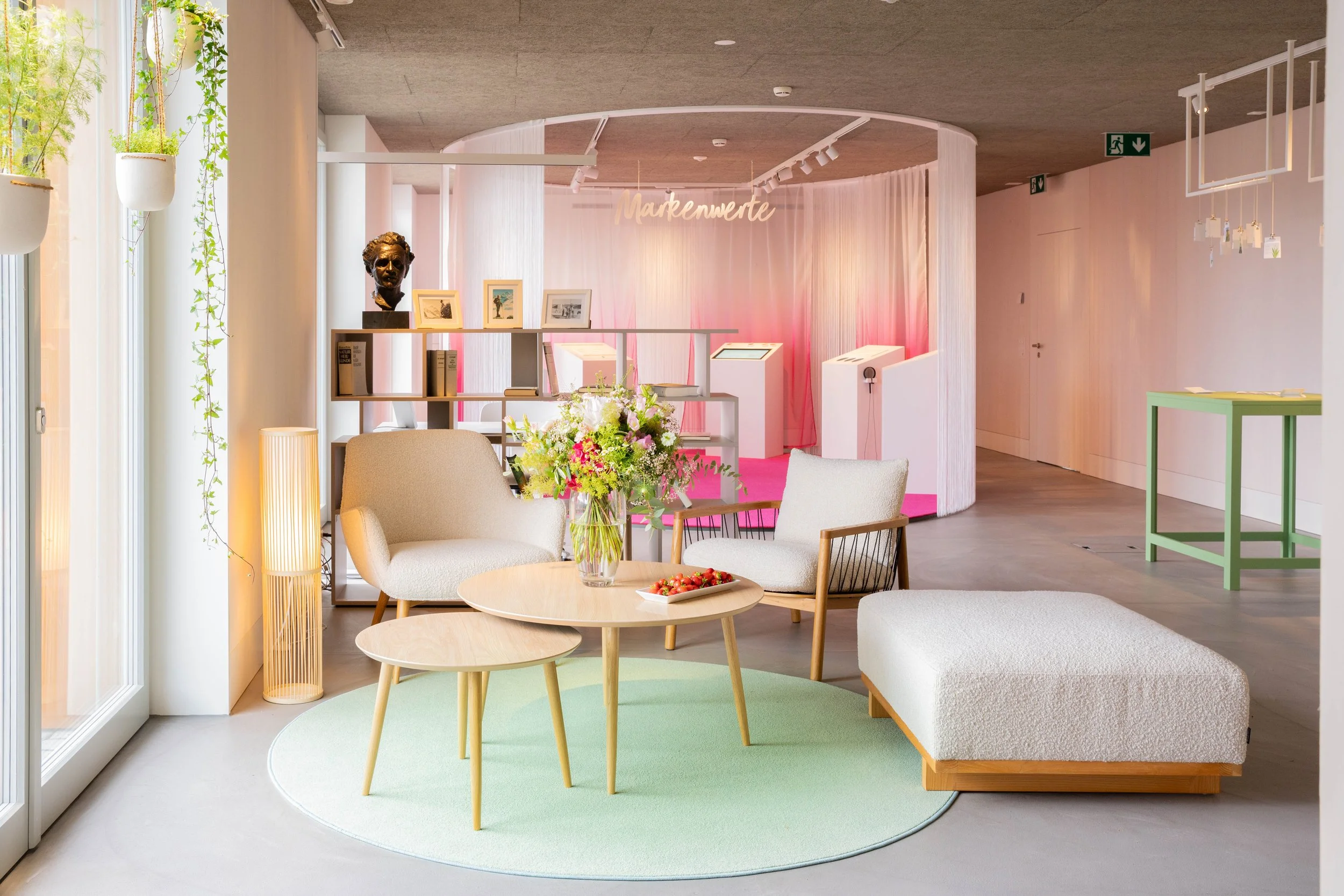 A cozy modern living space with beige armchairs, a white ottoman, and wooden coffee tables on a light green rug, decorated with a bouquet of colorful flowers and a plate of strawberries, next to large windows with hanging potted plants. In the background, there is an art or exhibit area with pink lighting and a sign that reads "Markenwerte."