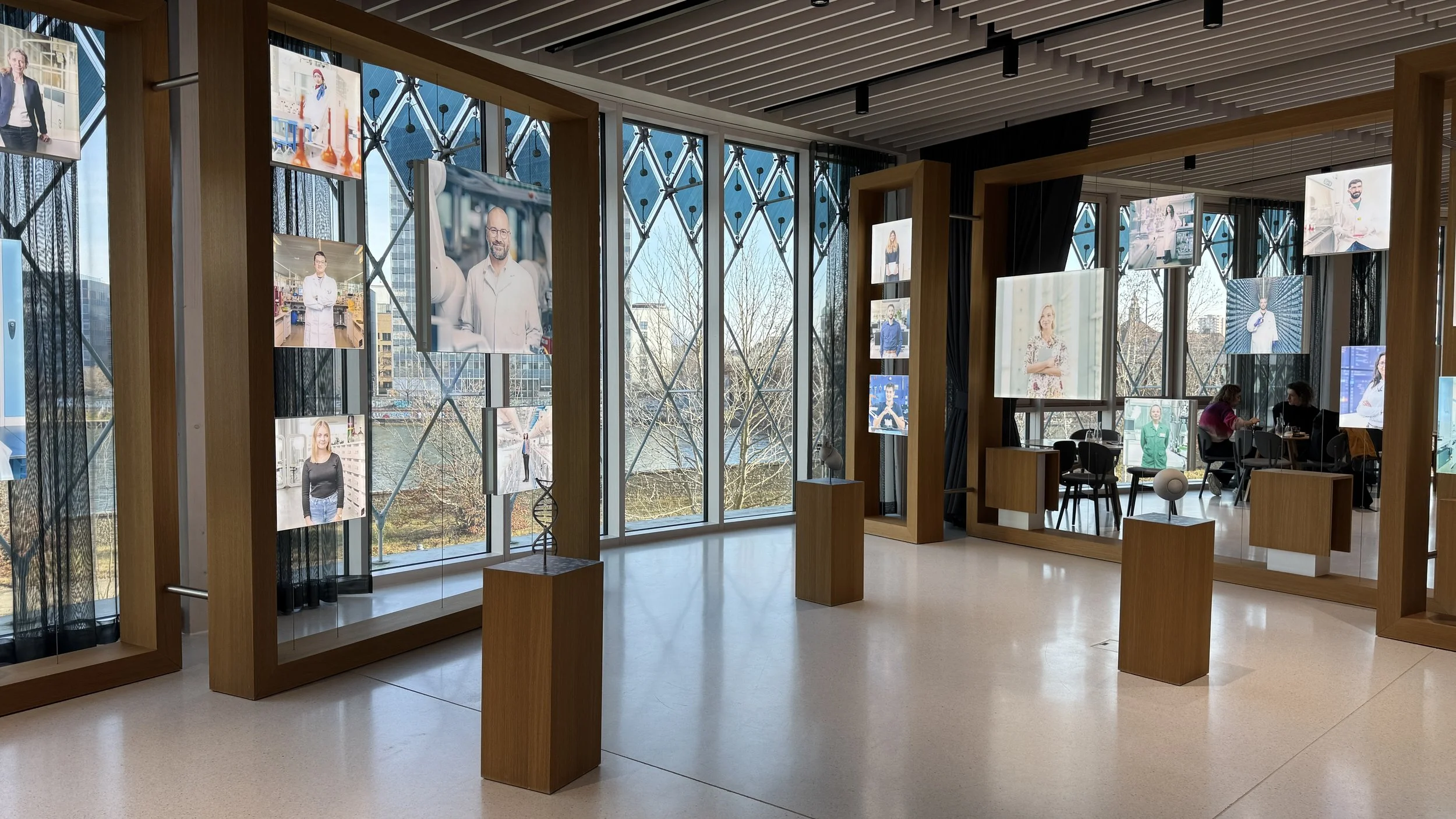 Indoor gallery with large floor-to-ceiling windows, hanging framed photographs of people, and a seating area with two individuals talking at a table.